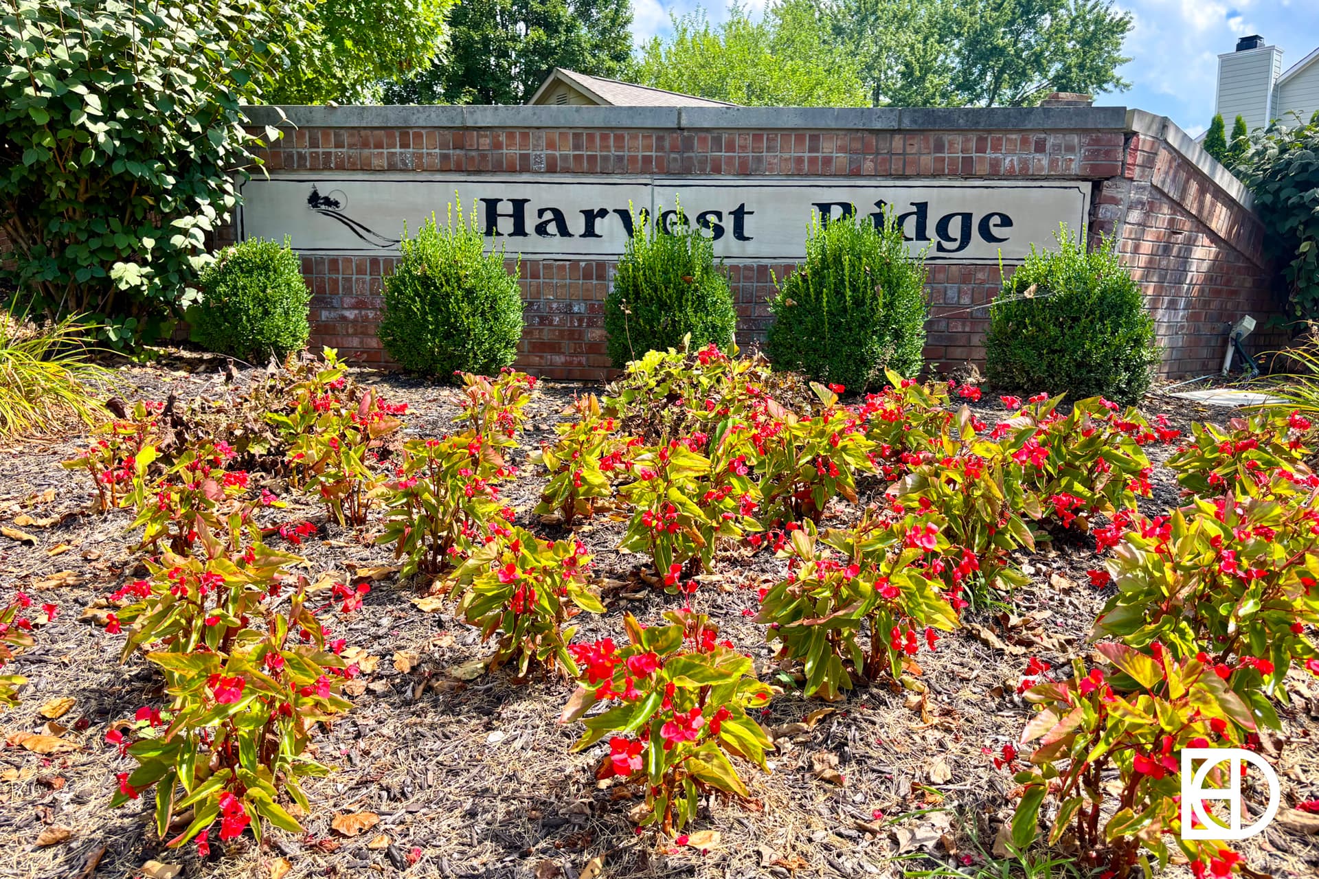 A brick sign reading Harvest Ridge is surrounded by green bushes and red flowers, with trees and a house visible in the background under a sunny sky.