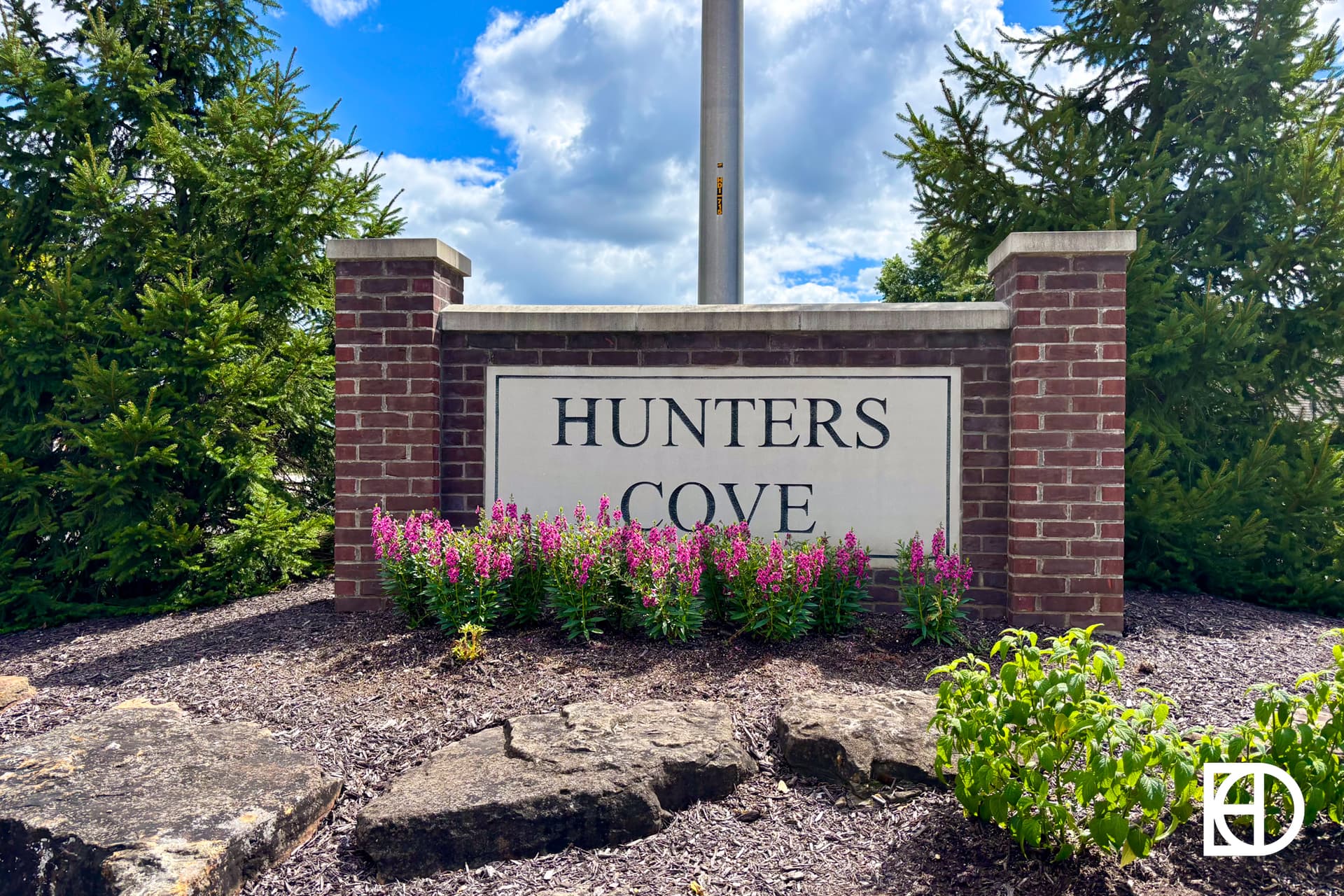 A brick sign with Hunters Cove written on it stands in front of green pine trees and purple flowers, under a partly cloudy blue sky.
