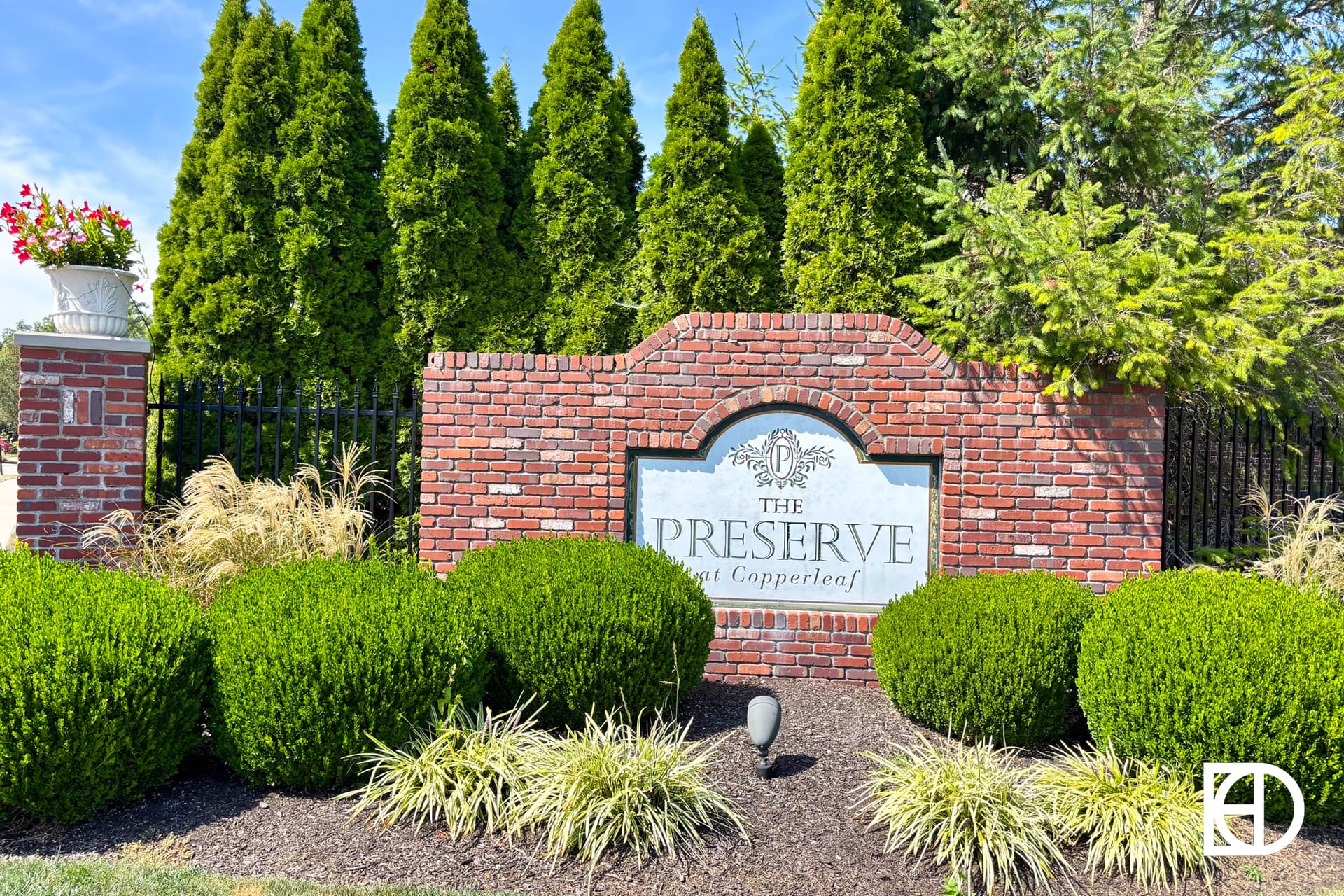 A brick sign reads The Preserve at Copperleaf, surrounded by neatly trimmed bushes, tall evergreen trees, and a planter with pink flowers, under a blue sky.