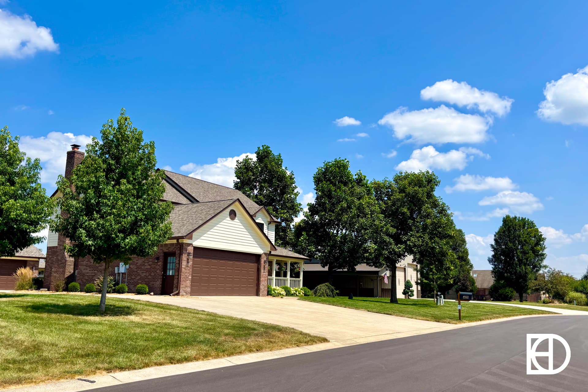 A quiet suburban street ends in a cul-de-sac lined with green trees, a single-story house, and a detached garage on a sunny day with blue sky and scattered clouds. A white KD logo appears in the bottom right corner.