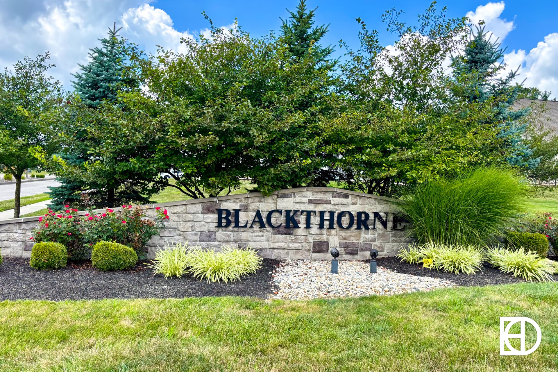 A stone sign reading BLACKTHORNE is surrounded by landscaped greenery, including bushes, ornamental grasses, and small trees, with a partly cloudy sky above.