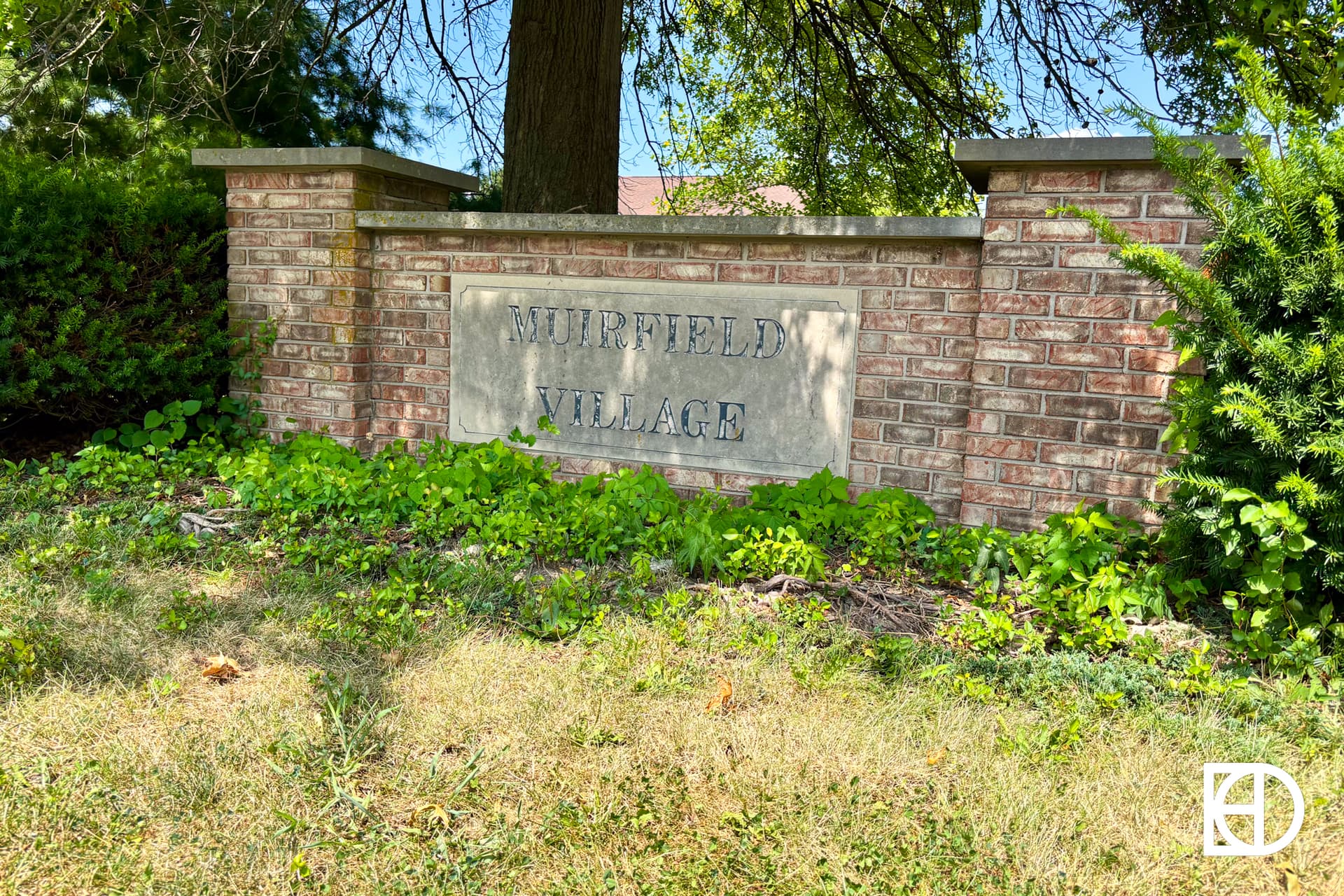 A brick sign reading Muirfield Village stands among green plants and grass, backed by trees and partially shaded.
