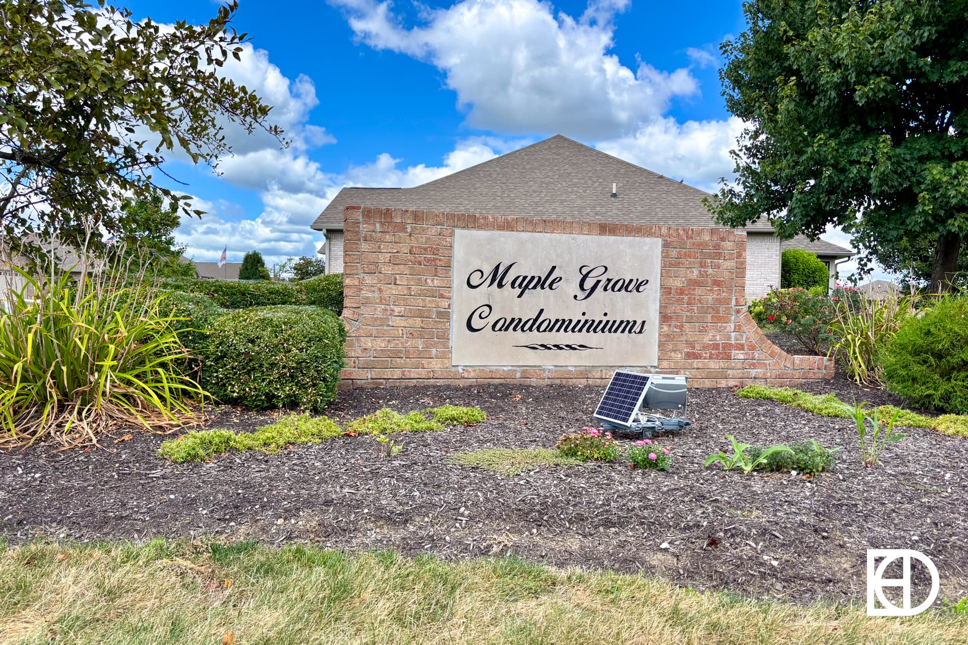 A brick sign reading Maple Grove Condominiums stands in a landscaped area with mulch, plants, and a small solar light in front. Houses and trees are visible in the background under a partly cloudy sky.