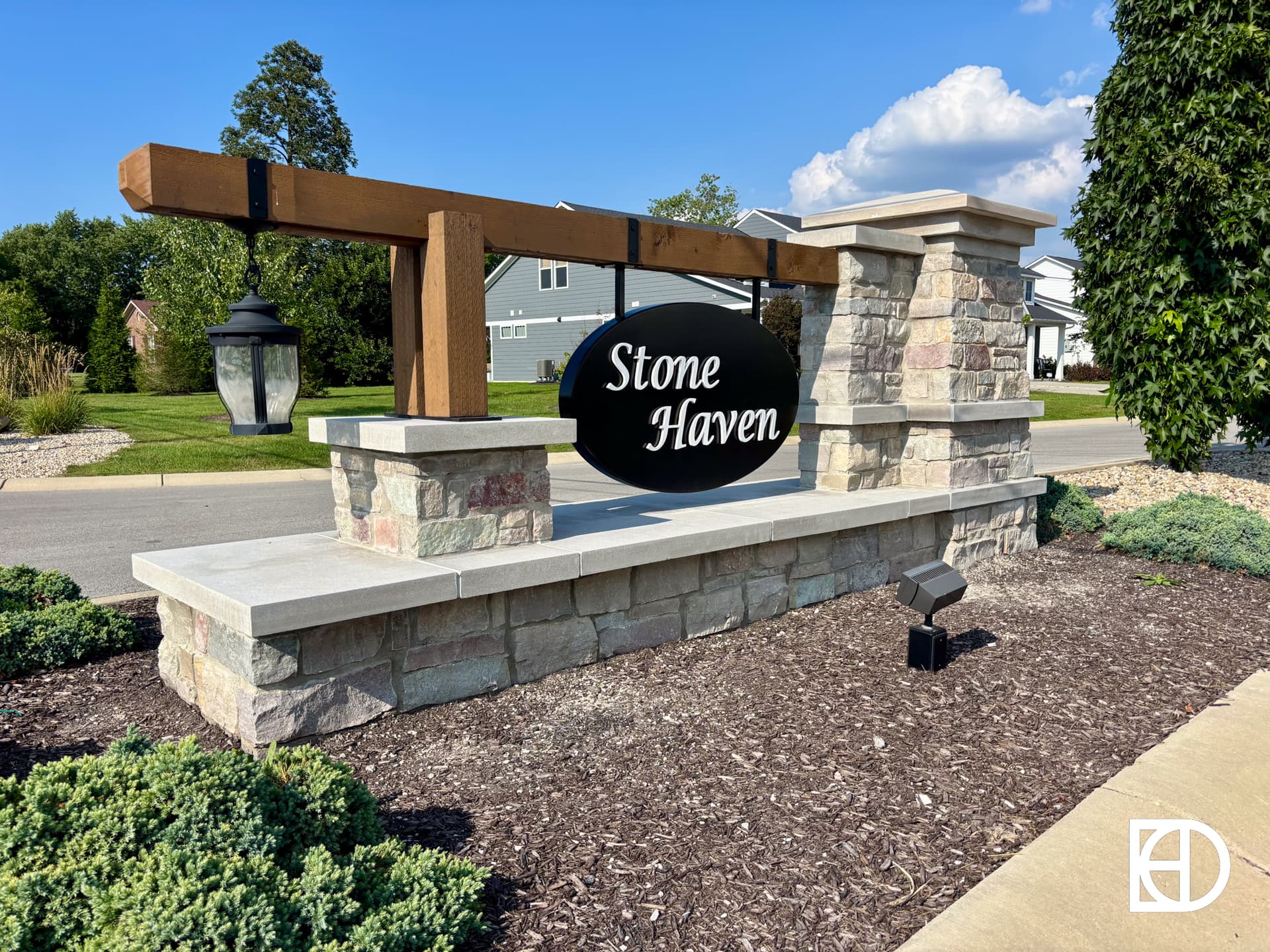 A stone and wood entrance sign reads Stone Haven, with a large black oval sign hanging from a wooden beam and a lantern, set near a road and surrounded by landscaped bushes.