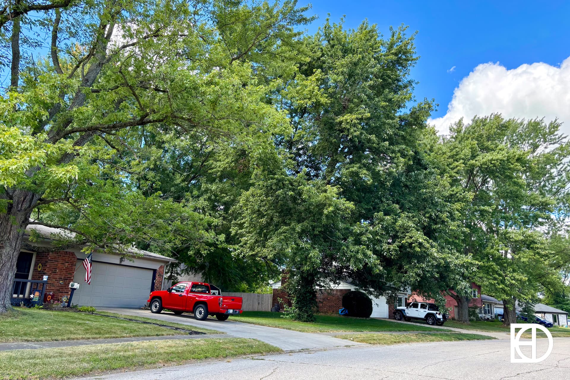 A suburban street with houses partially hidden by large green trees, a red pickup truck parked in a driveway, and an American flag hanging near a garage. The sky is mostly blue with a few clouds.