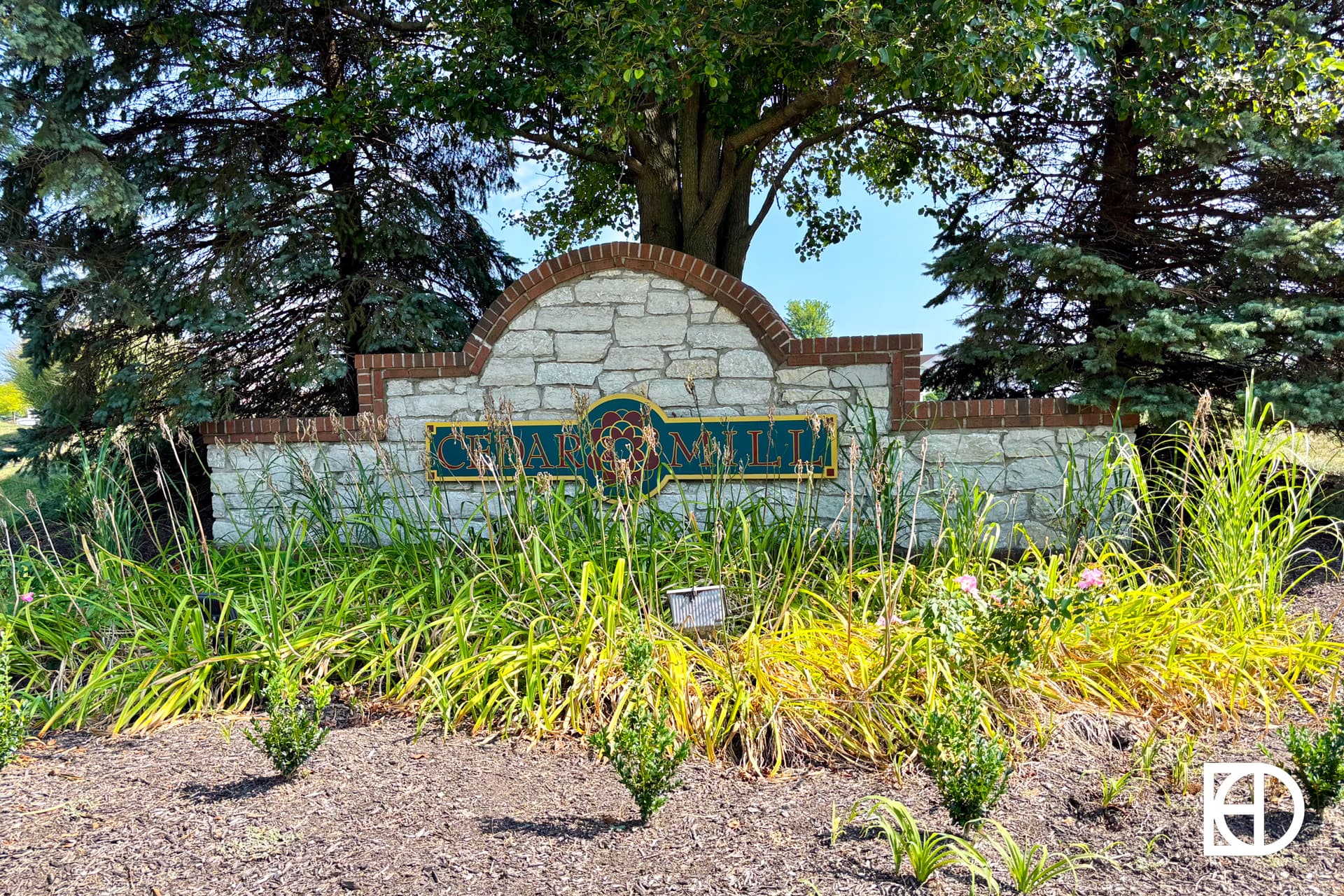 A stone and brick entrance sign with “Quarry Mill” written on it, surrounded by green plants, flowers, and trees in the background, on a sunny day.