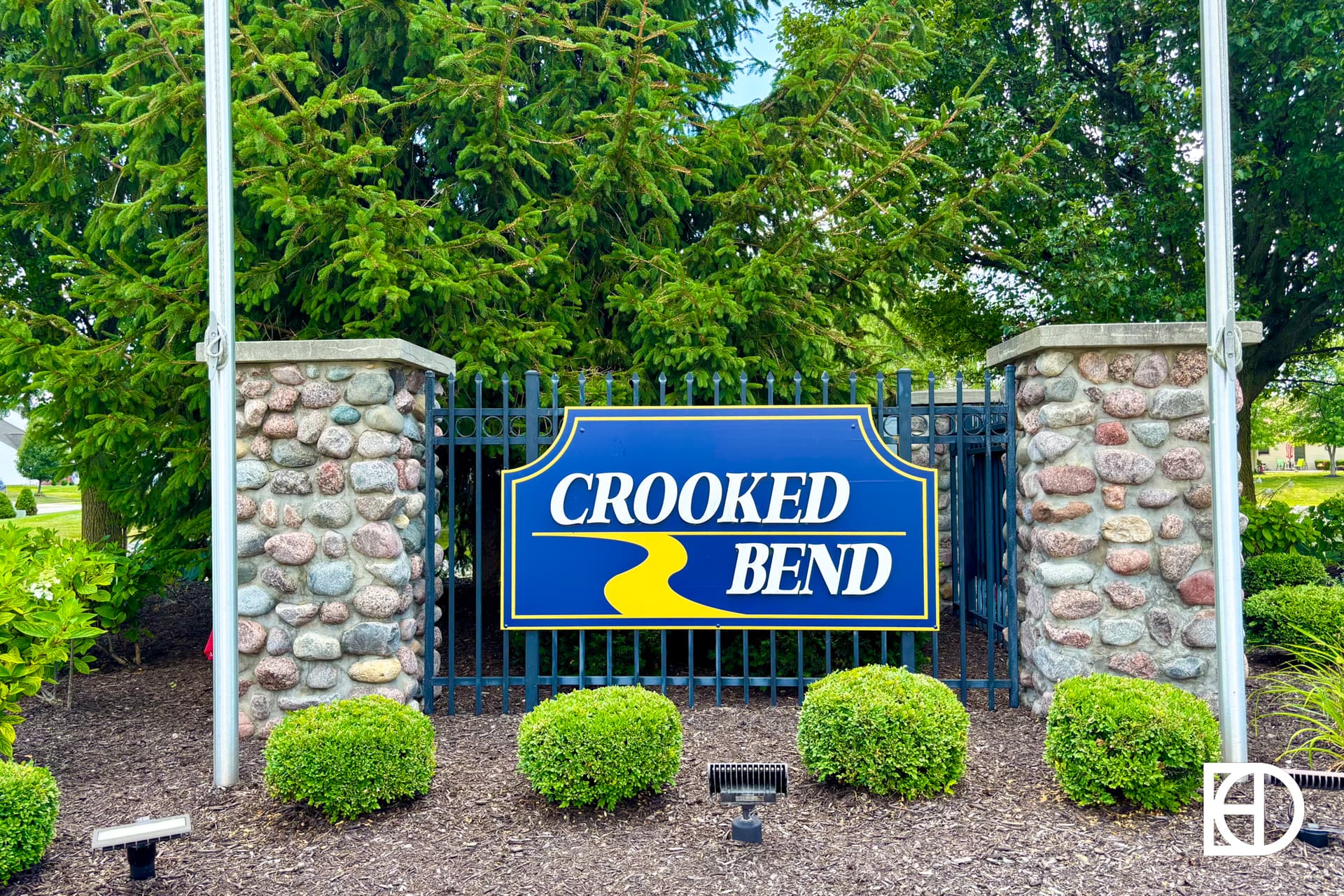 A stone and metal sign reads Crooked Bend in yellow and white letters on blue, with a yellow curved line below, surrounded by greenery and shrubs.