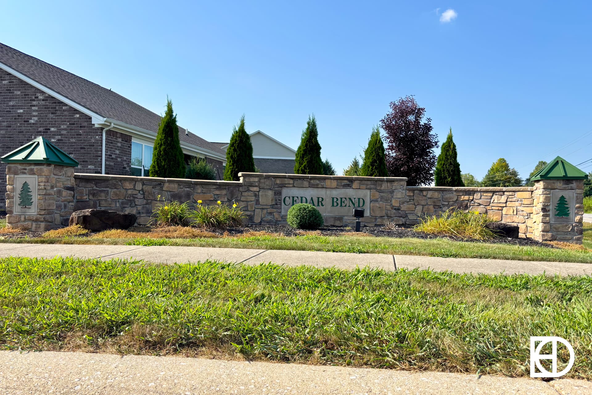 A stone entrance sign with green accents reads Cedar Bend. It is set in front of a landscaped area with small trees and shrubs, next to a residential building and sidewalk under a clear blue sky.