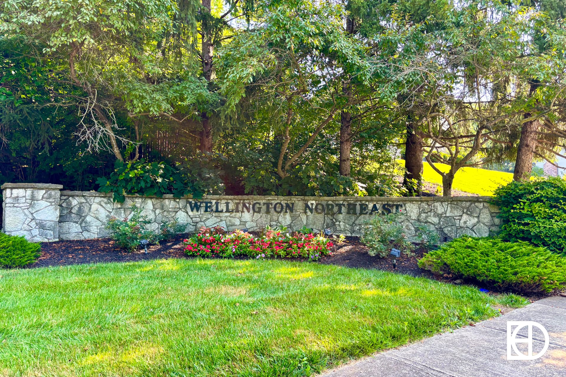 A stone wall with the words WELLINGTON NORTHEAST stands behind neatly trimmed grass, vibrant flowers, and bushes, surrounded by tall green trees on a sunny day.