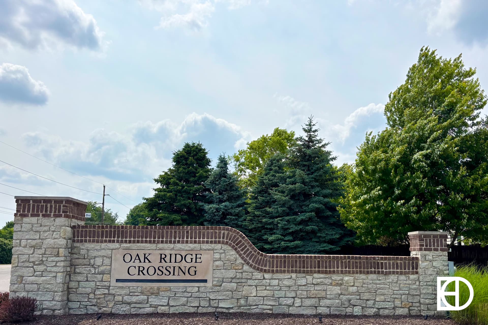 A brick and stone entrance sign reads Oak Ridge Crossing, surrounded by trees and a partly cloudy sky in the background.
