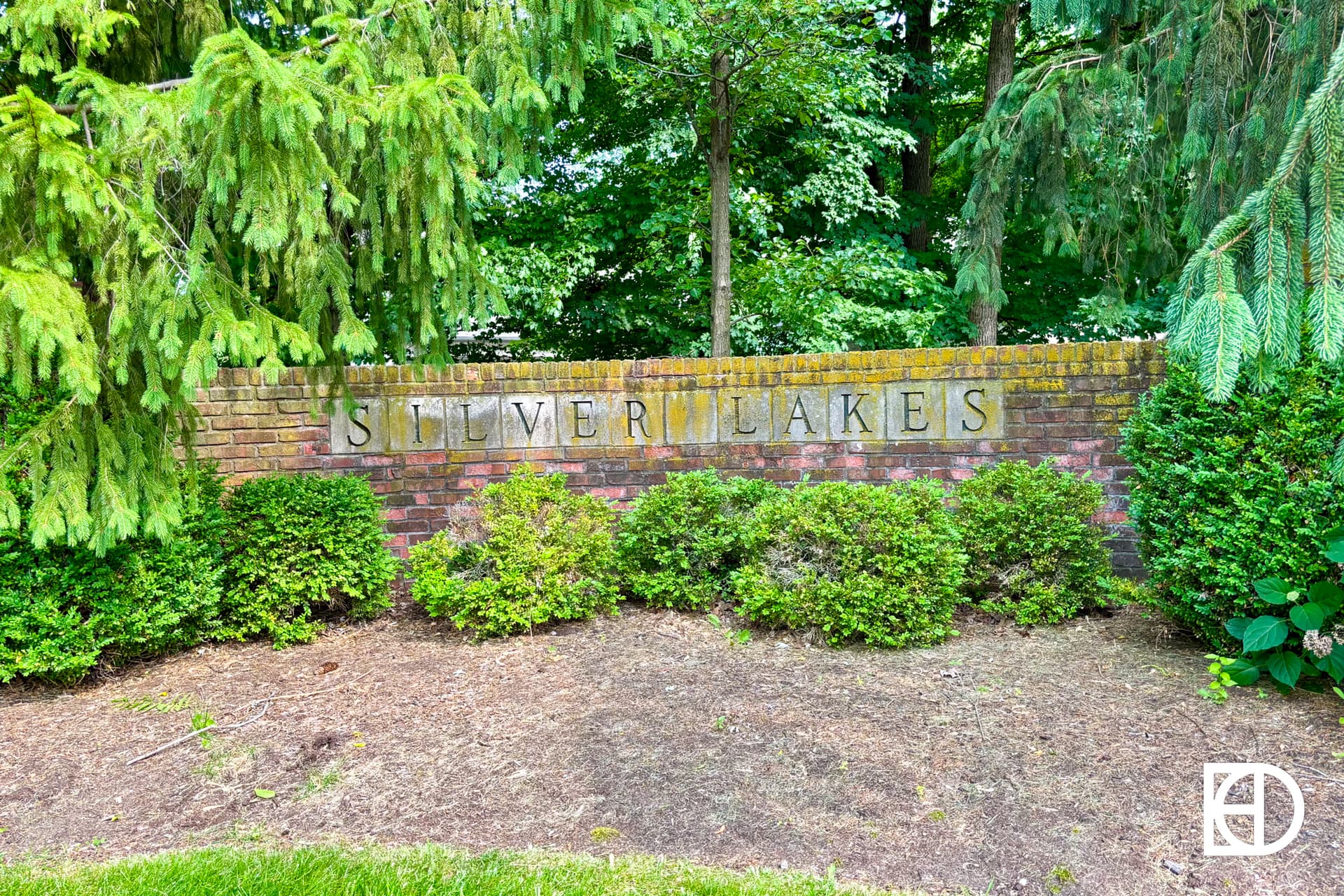 A brick wall with the words SILVER LAKES in block letters, surrounded by green shrubs and pine trees.