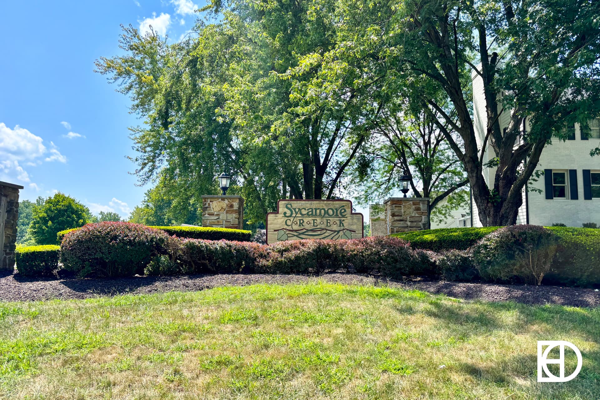 A stone sign reading Sycamore Creek sits among trimmed bushes and trees on a sunny day, with a large white building partially visible on the right.