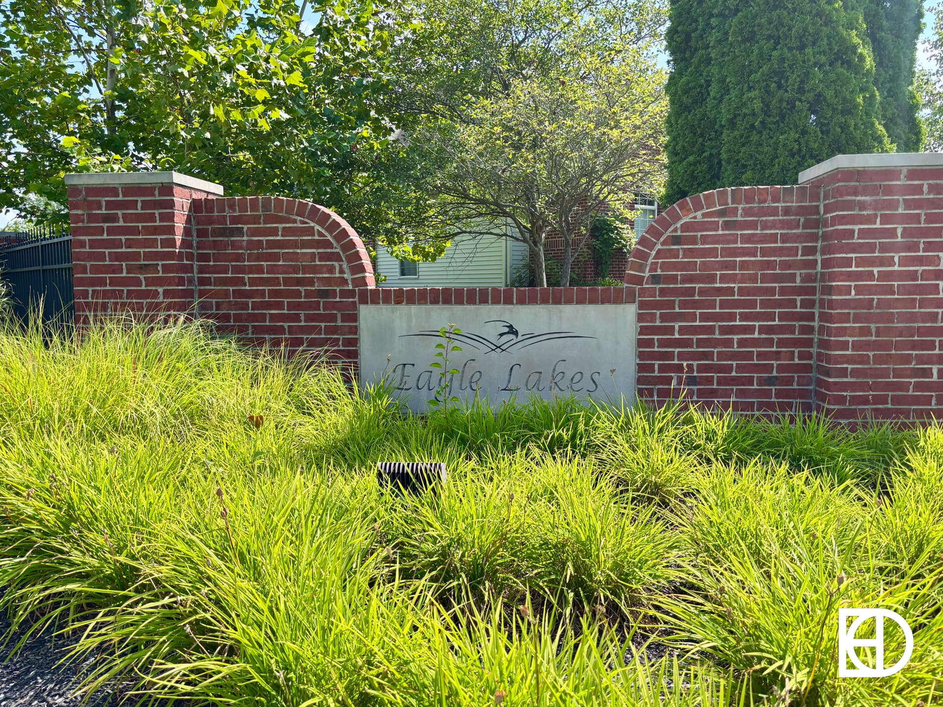 A brick entrance sign with “Eagle Lakes” written on a stone panel, surrounded by green grass and trees, under a clear sky.