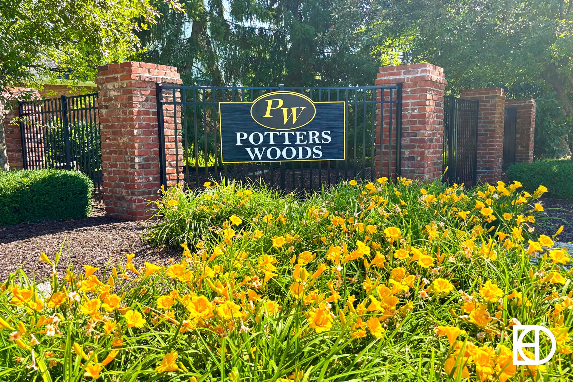 A brick entrance sign reading PW Potters Woods stands behind a black metal fence, surrounded by bright yellow flowers and lush green bushes on a sunny day.