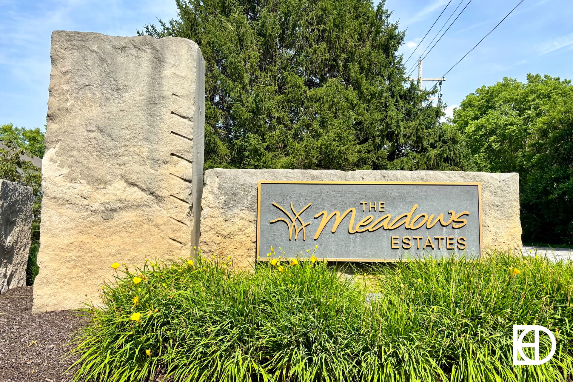 Large stone entrance sign with The Meadows Estates in cursive gold letters, surrounded by green bushes and yellow flowers, set against trees and a blue sky.