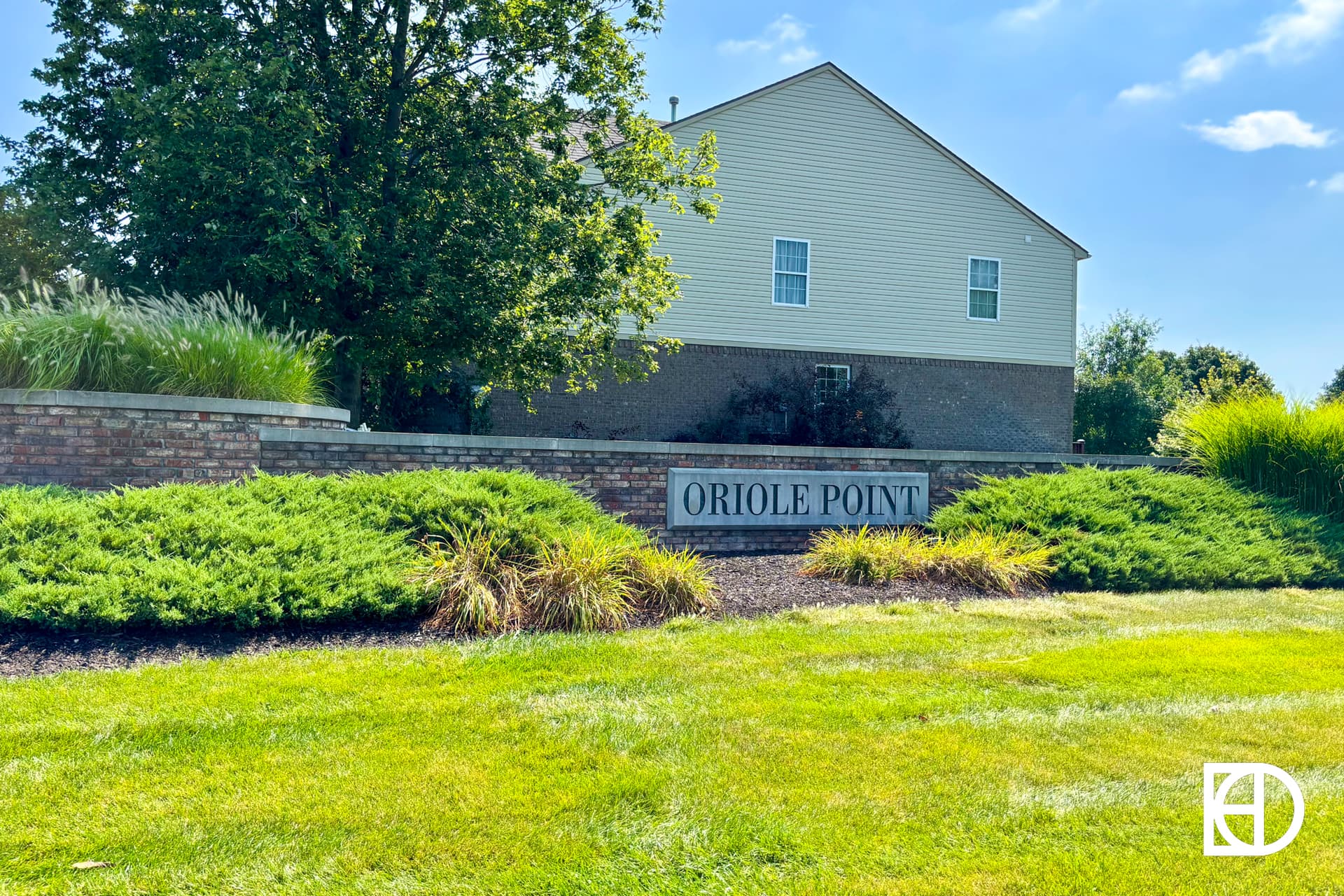 A brick entrance sign for Oriole Point stands amid green bushes and grass, with a two-story house and trees in the background under a blue sky.