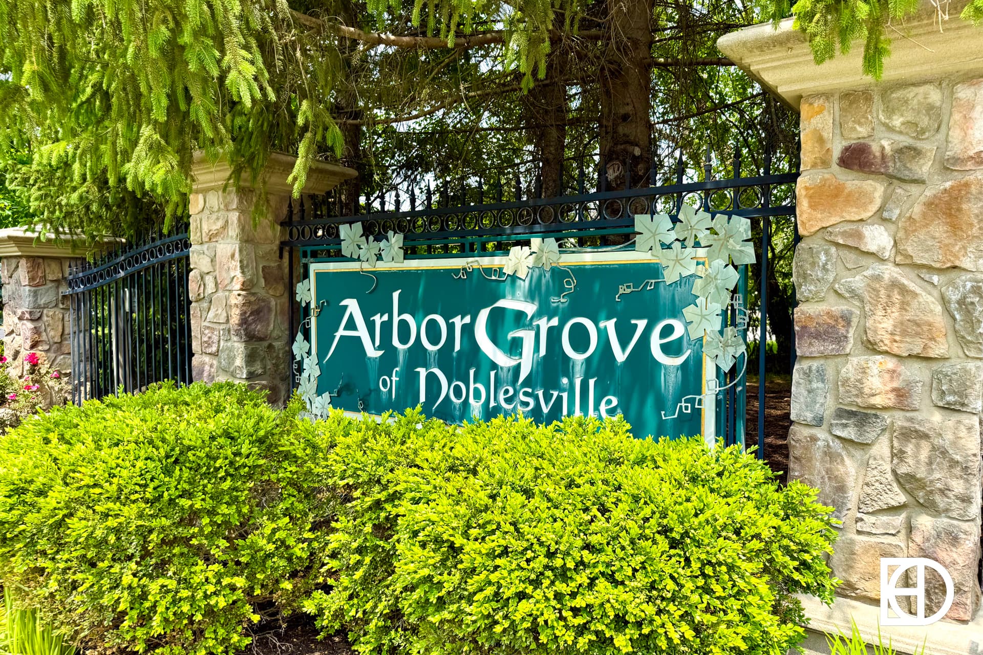 A green and white sign reading Arbor Grove of Noblesville is mounted on a black fence, surrounded by stone pillars, bushes, and trees.