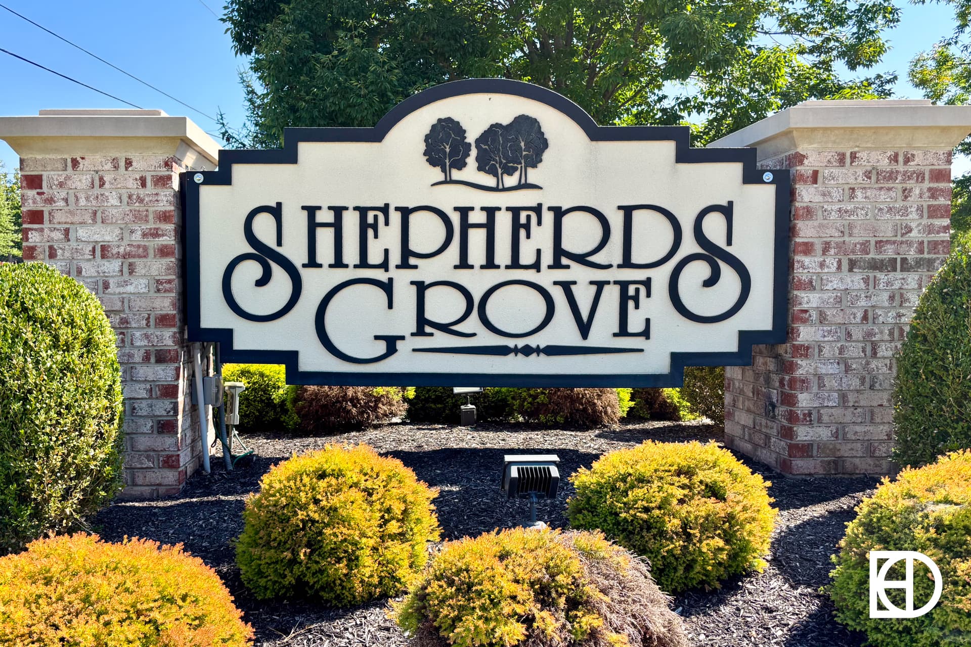 A decorative sign reading Shepherds Grove is mounted between two brick pillars, surrounded by neatly trimmed shrubs and greenery, with a blue sky and trees in the background.