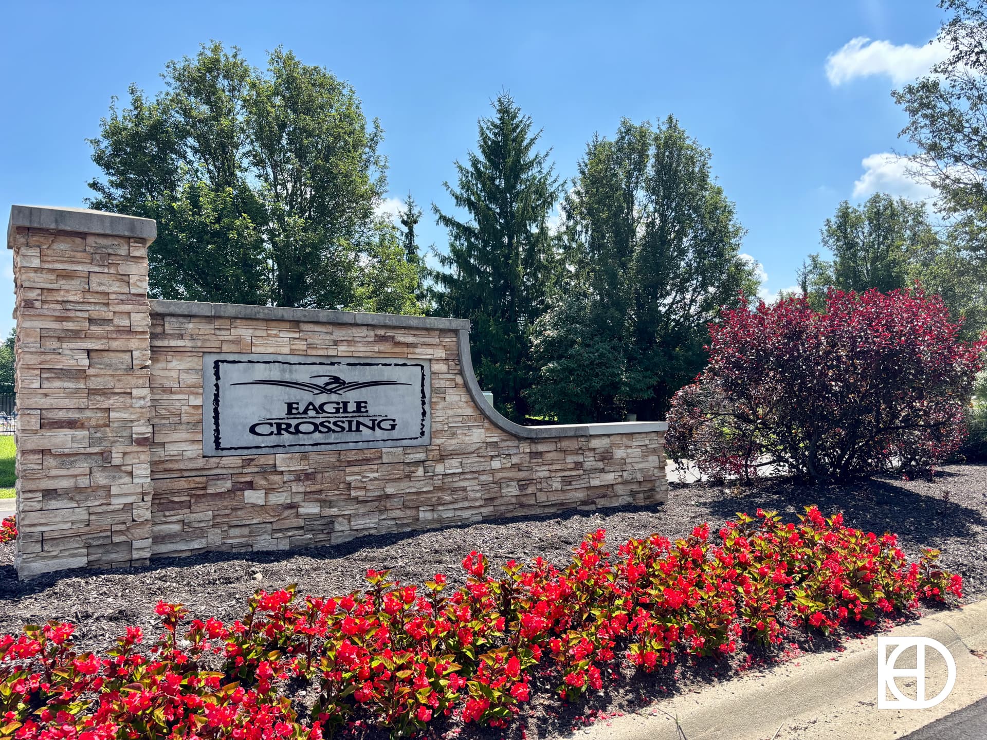 A stone entrance sign reads Eagle Crossing with an eagle image, surrounded by red flowers, shrubs, and trees under a blue sky.