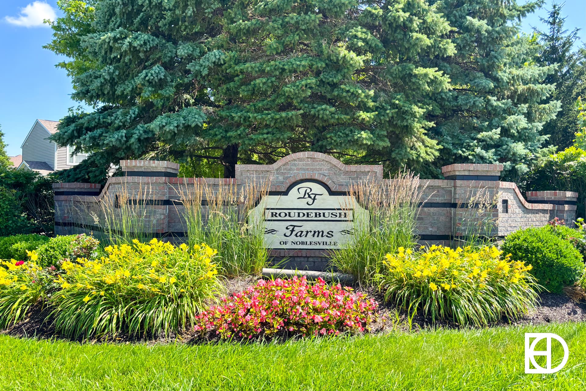 A landscaped stone sign reads Roudebush Farms of Noblesville, surrounded by green bushes, yellow and pink flowers, and tall grass, with trees and a clear blue sky in the background.