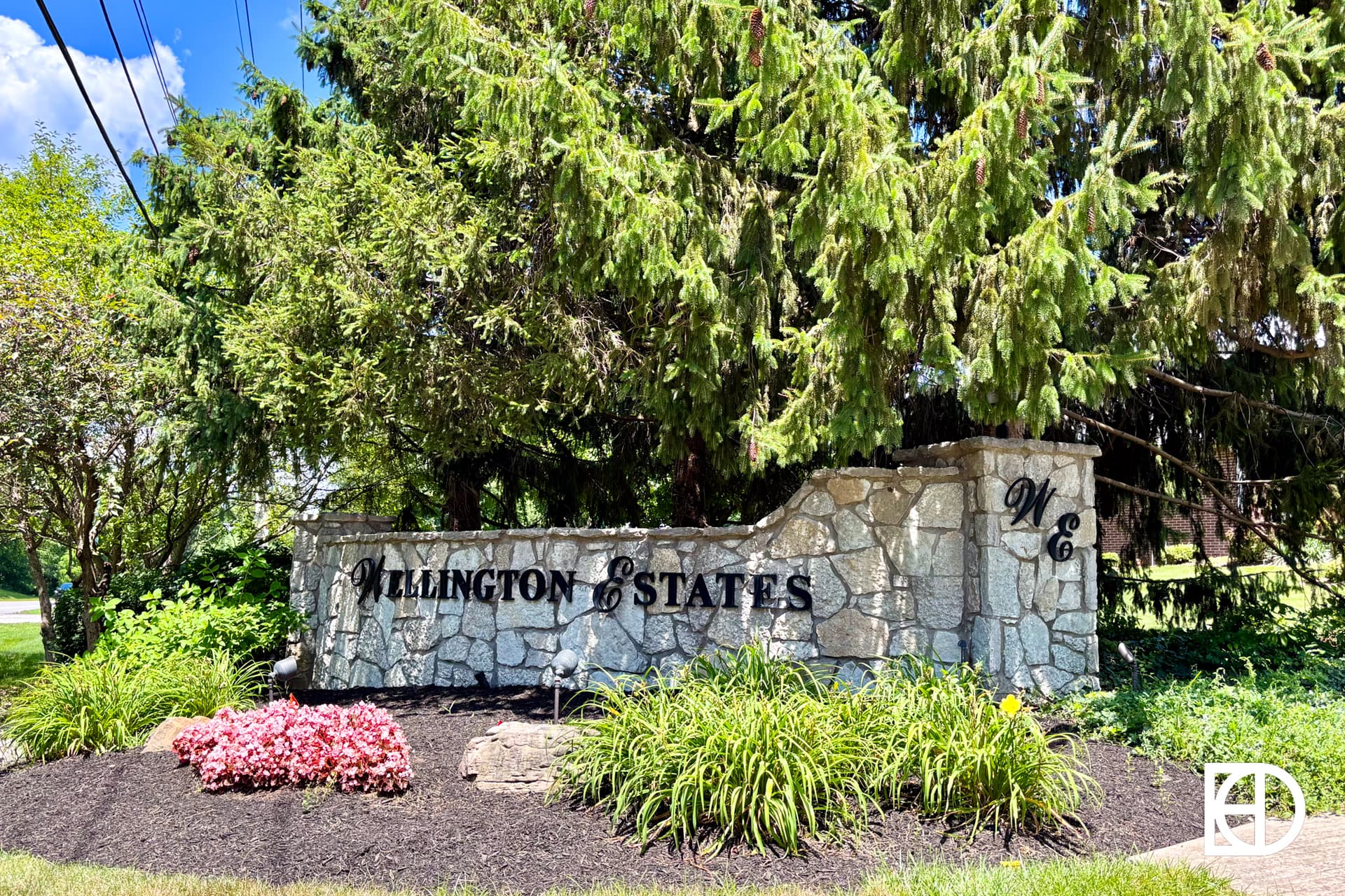 A stone entrance sign reading Wellington Estates with the letters WE, surrounded by green bushes, grass, and pink flowers, stands beneath a large tree on a sunny day.