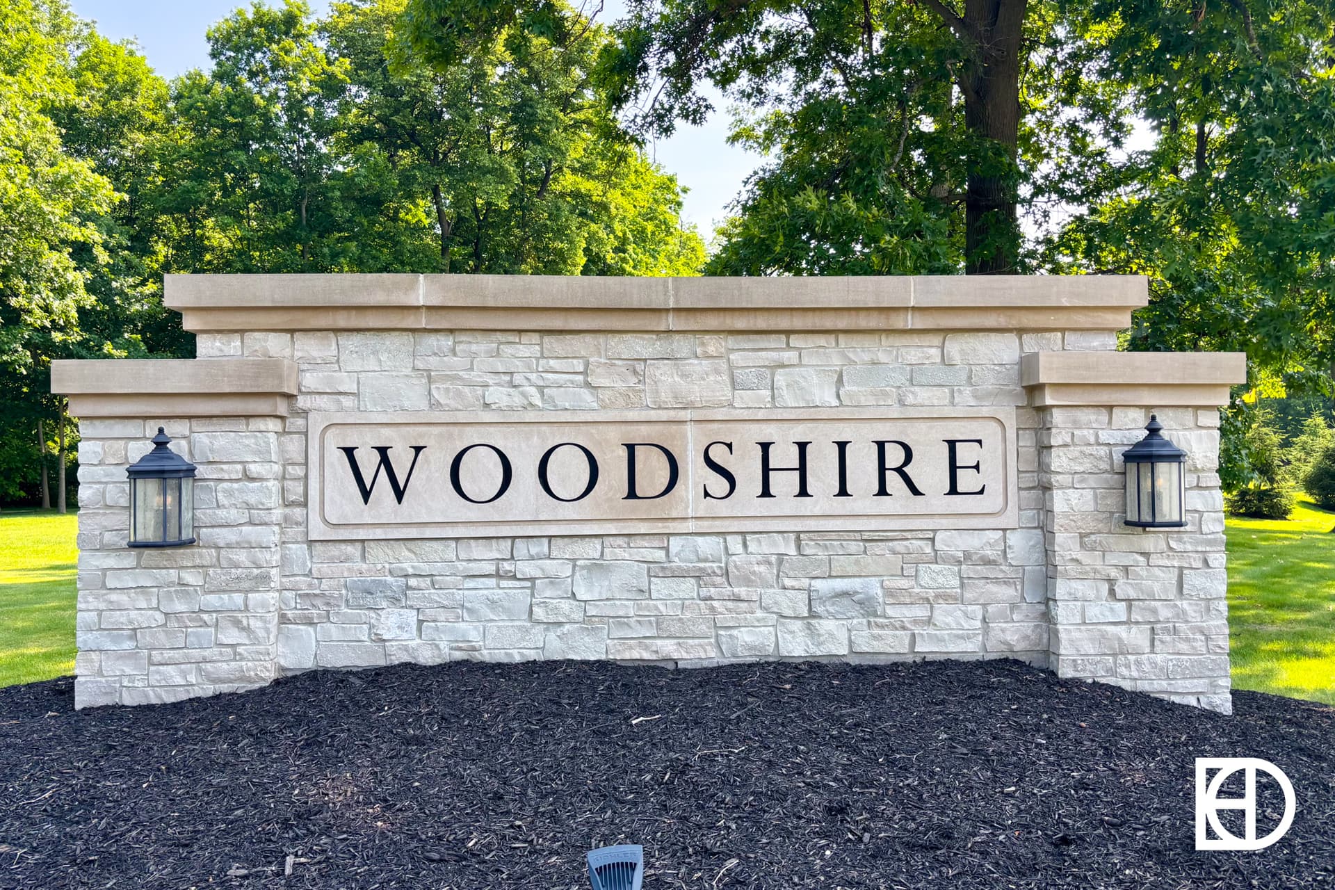 A large stone entrance sign with the word WOODSHIRE in black letters, flanked by two lantern-style lights, stands in front of green trees and grass under a clear sky.