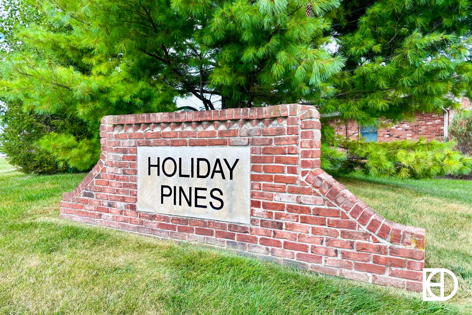 A brick sign with a plaque reading HOLIDAY PINES stands on a grassy area, surrounded by green pine trees. A white logo is visible in the bottom right corner of the image.