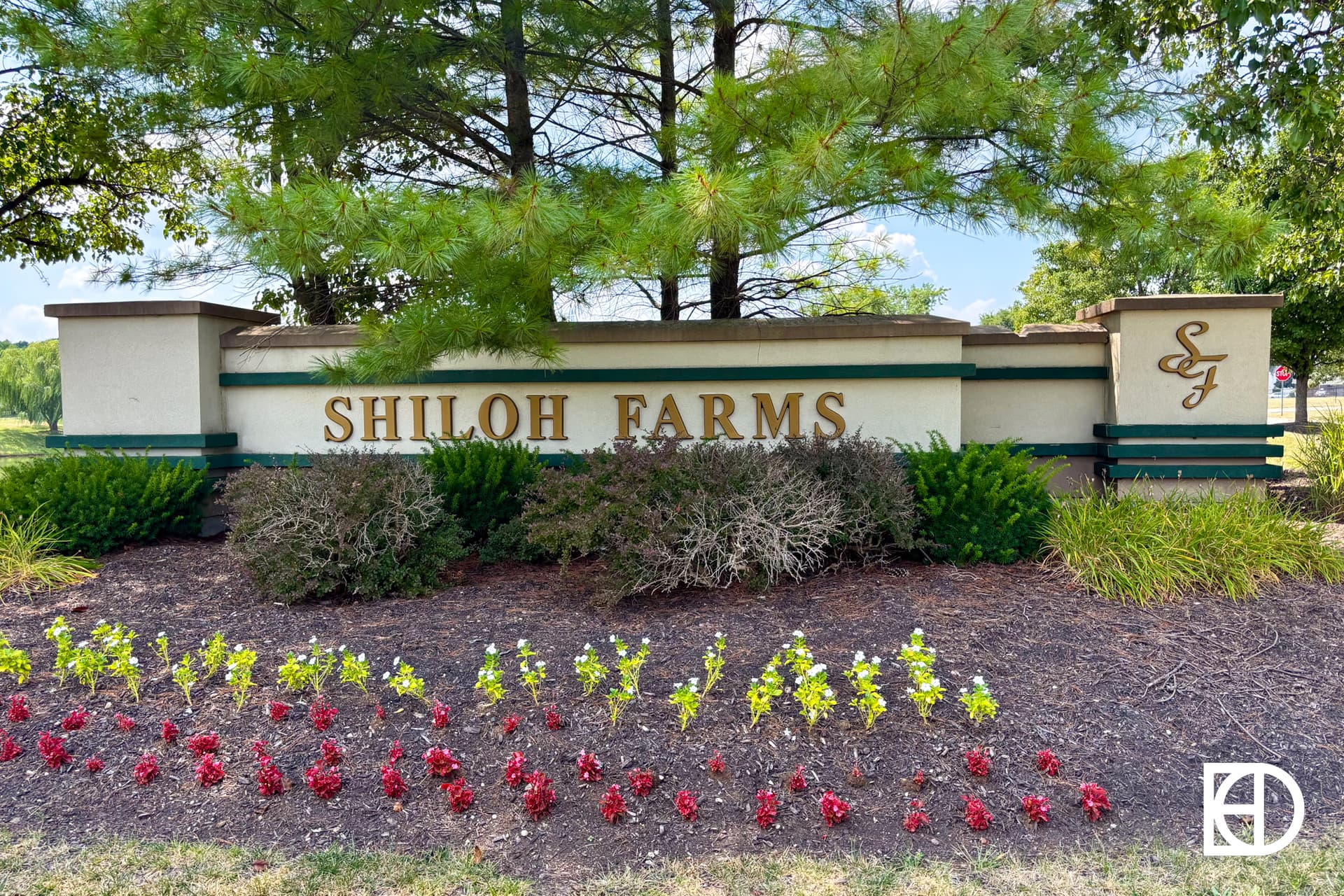 A stone entrance sign with SHILOH FARMS in gold letters, surrounded by bushes and colorful flowers, sits beneath tall pine trees on a sunny day. A decorative SF monogram is on the right side of the sign.