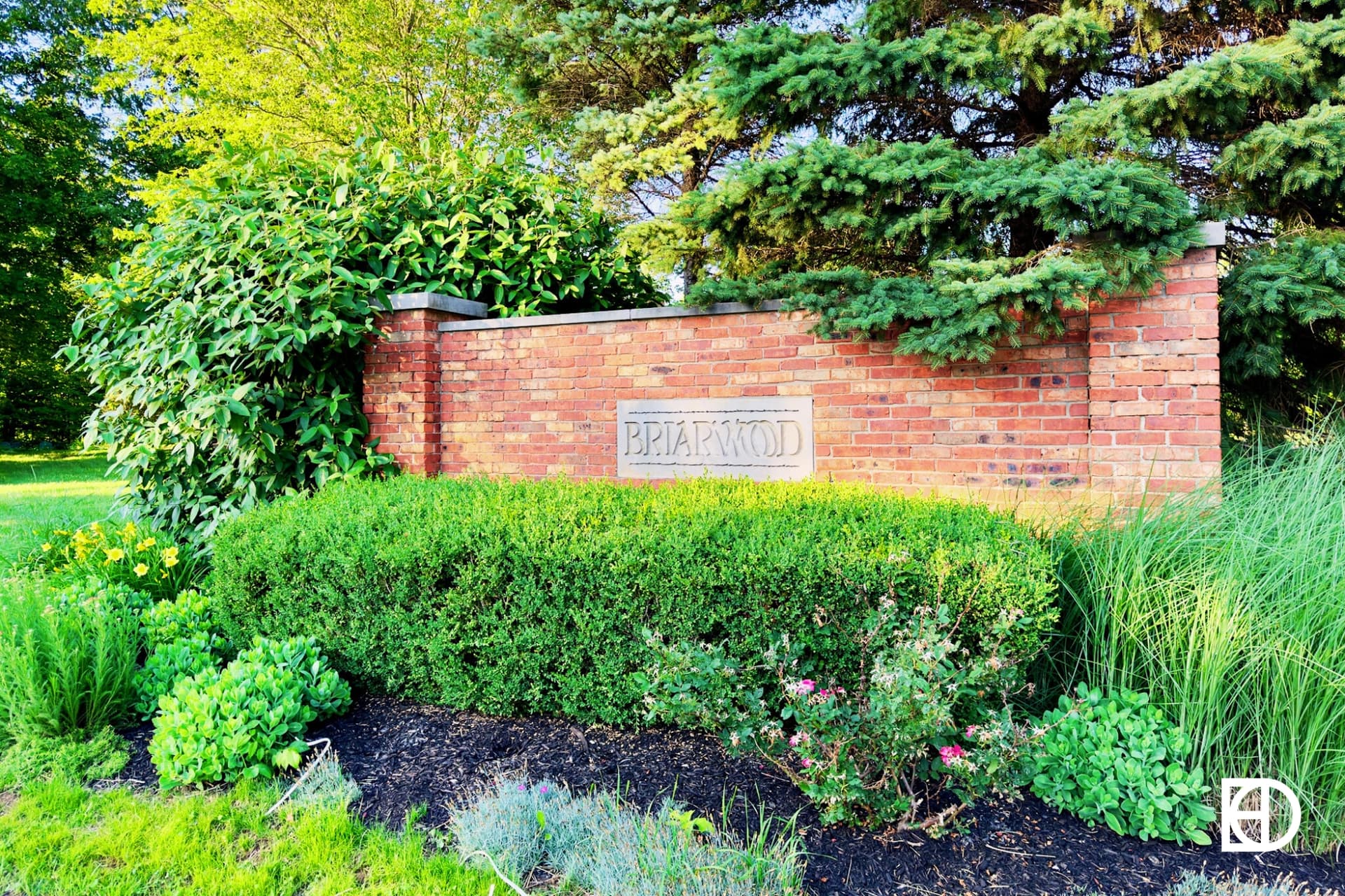 Photo of brick entrance sign with shrubs and flowers in front and trees behind.