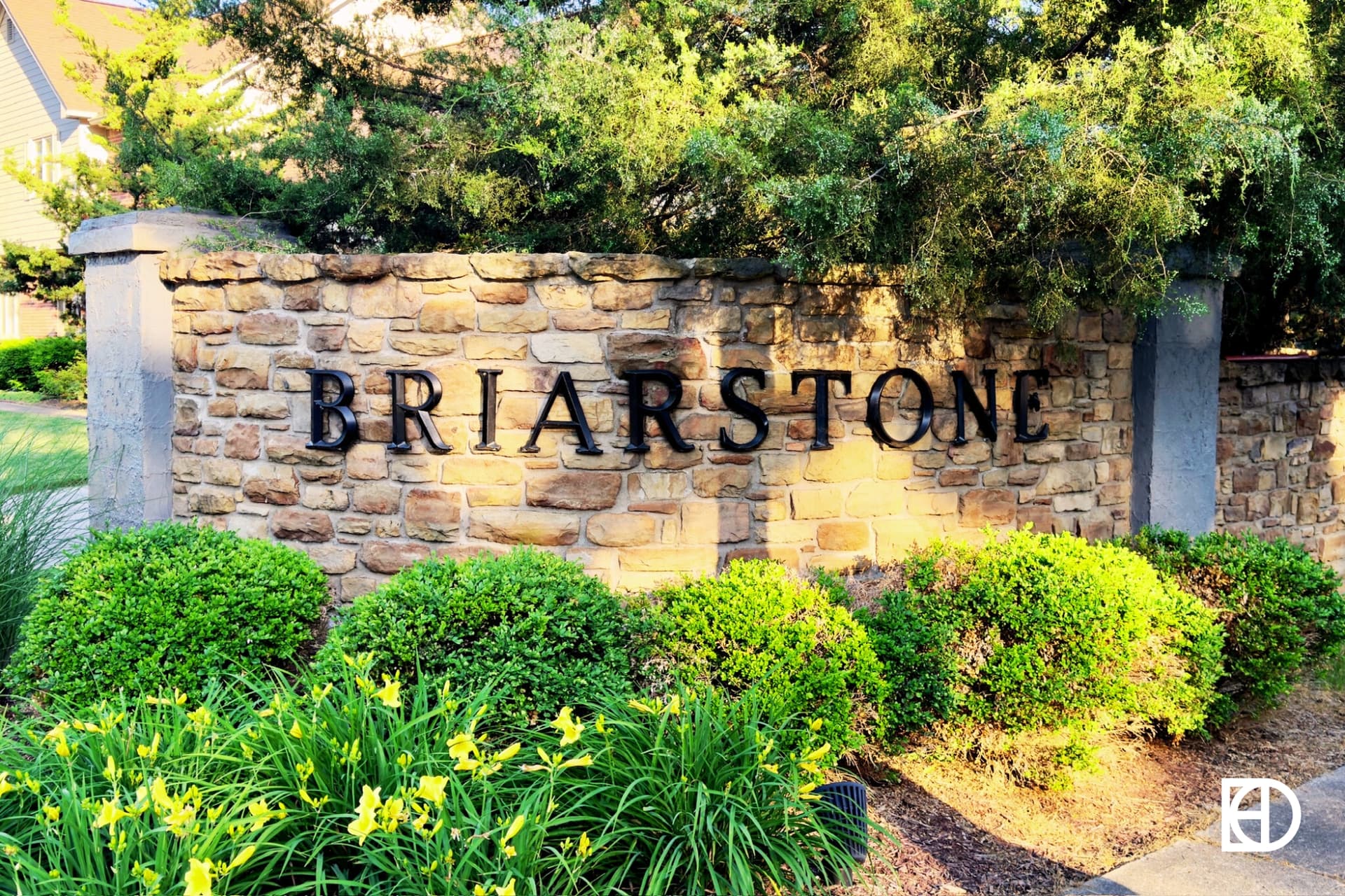 Photo of brick and stone entrance sign flanked by shrubs, daylilies and trees with the name Briarstone in black letters.