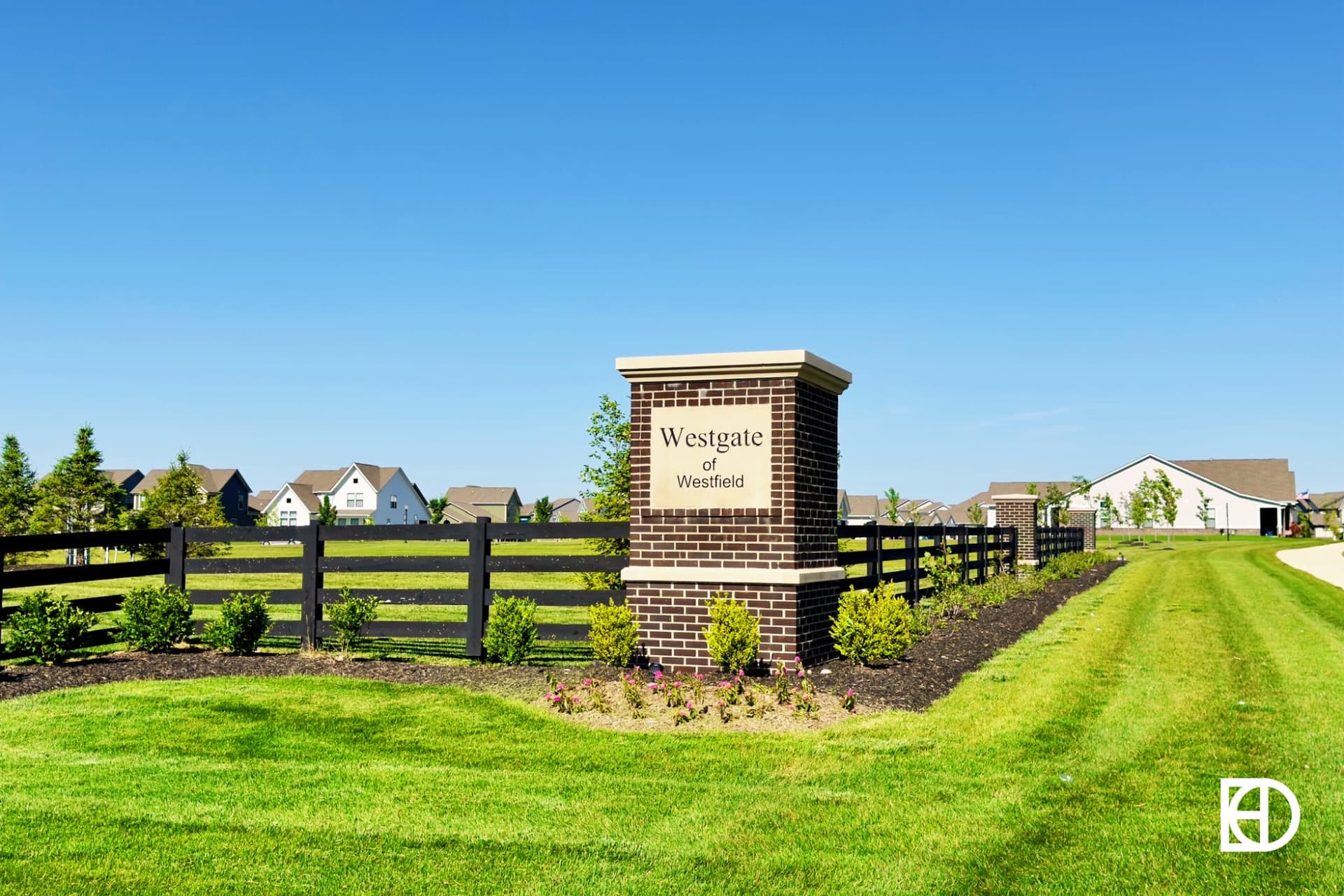 Brick entrance sign reading Westgate of Westfield stands by a black fence, with green lawns and suburban houses visible in the background under a clear blue sky.