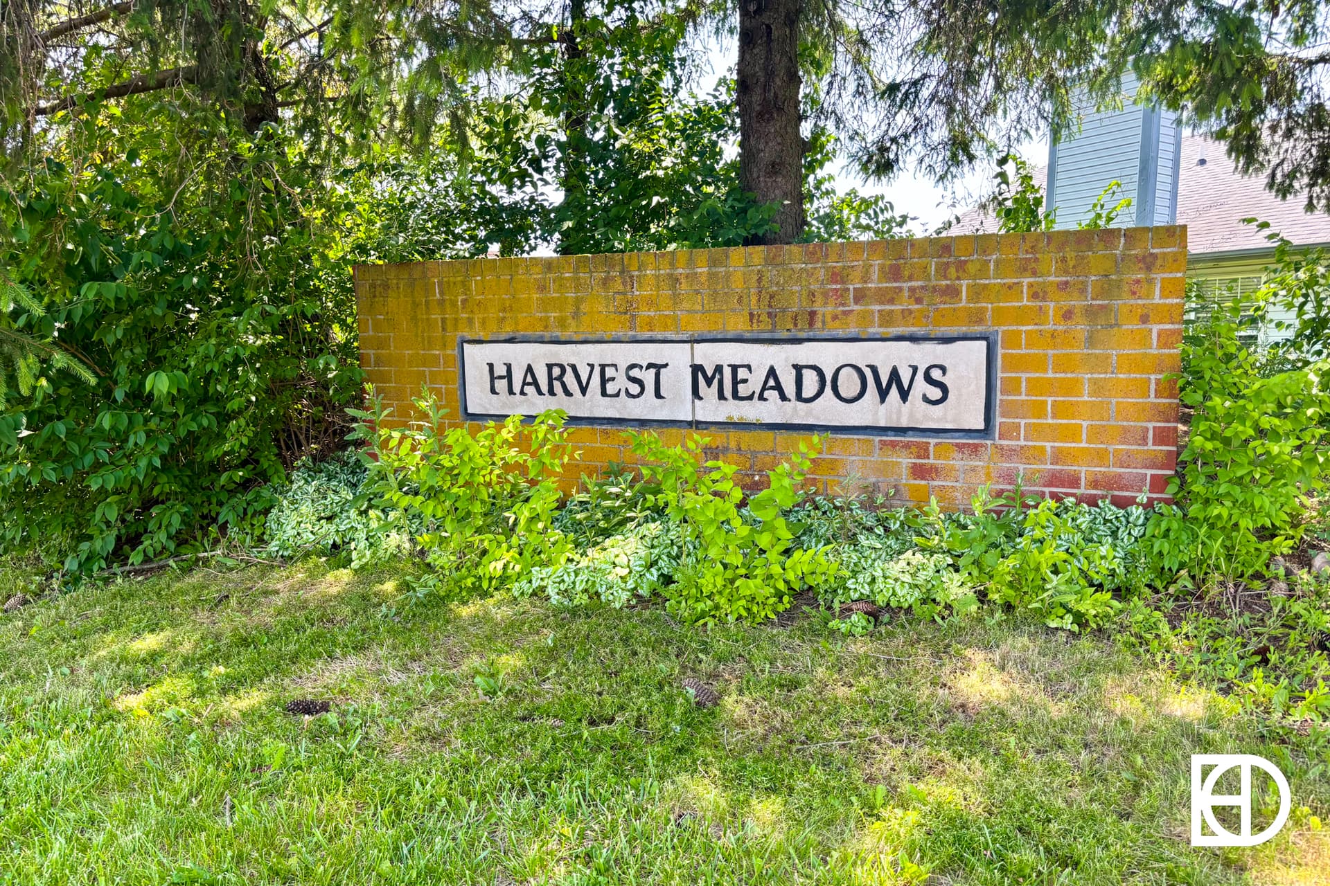 A brick sign reading Harvest Meadows is surrounded by green plants and trees on a grassy lawn.