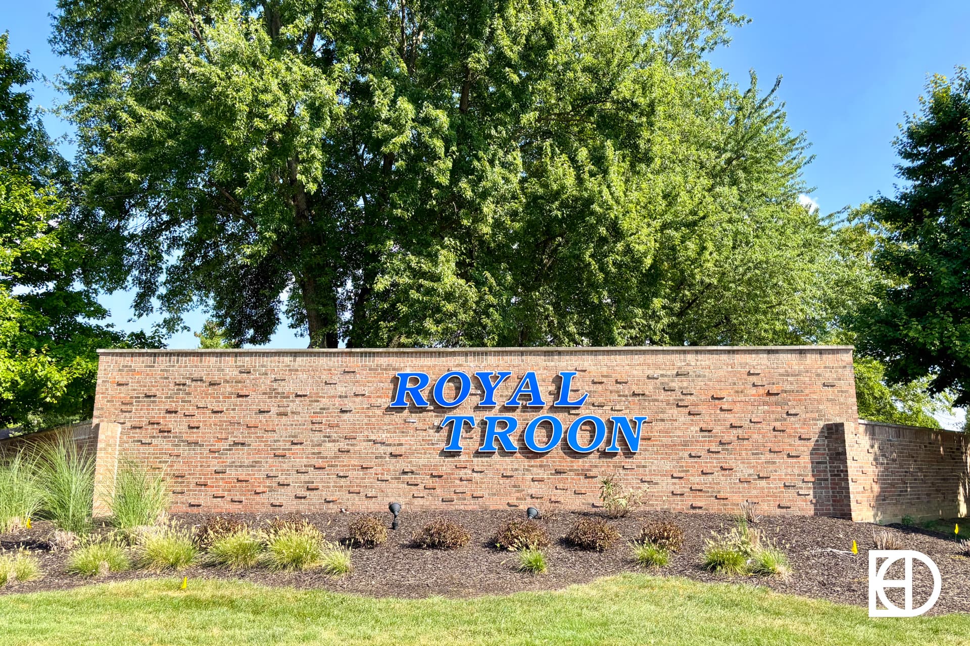 A brick sign with blue letters spelling Royal Troon stands in front of tall green trees and landscaping with small bushes and mulch.