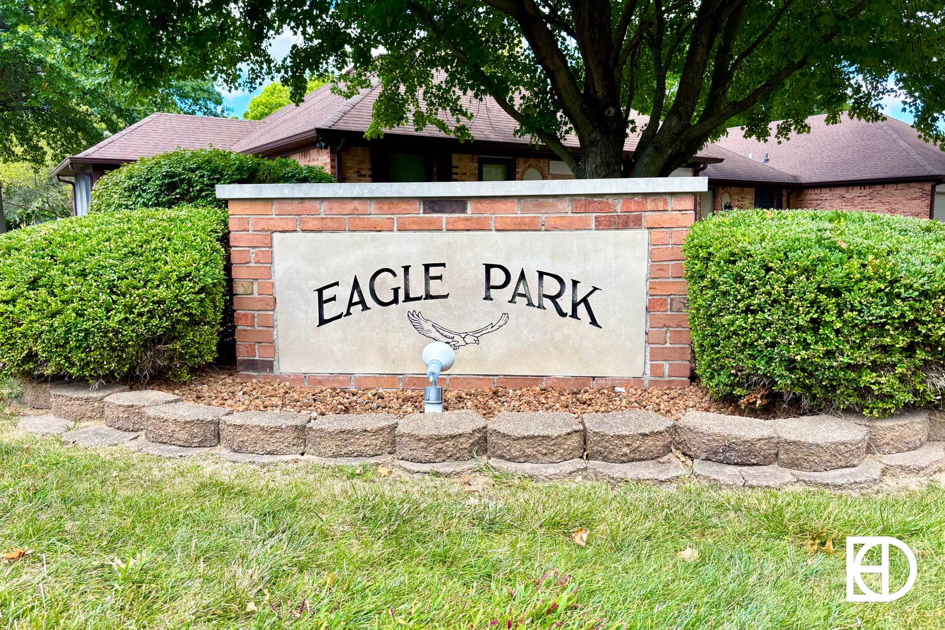 A brick and stone sign reading Eagle Park with an eagle illustration, surrounded by neatly trimmed bushes and a grassy area, in front of a house and shaded by a large tree.