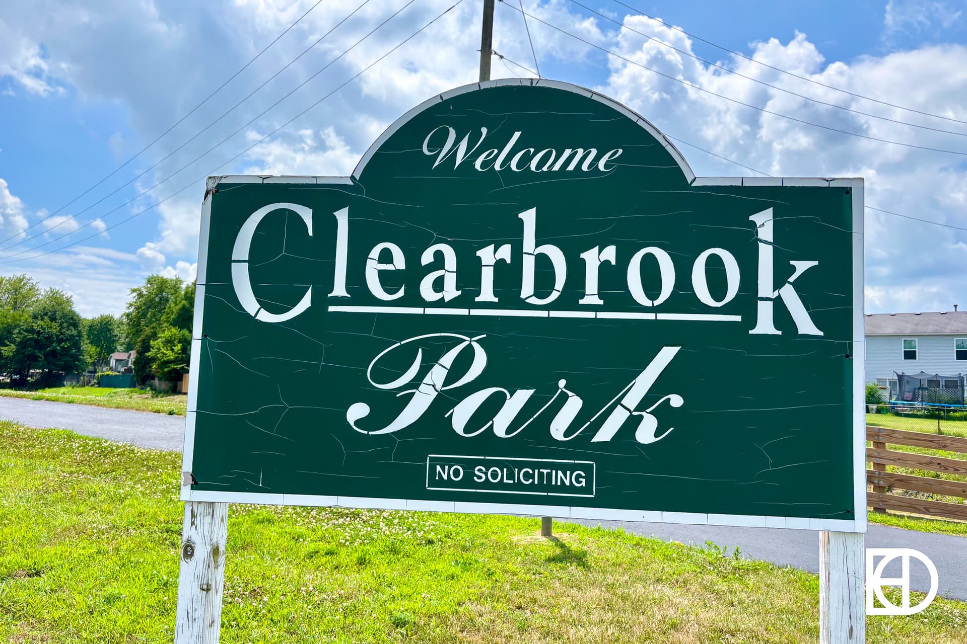 A dark green sign with white text reads Welcome Clearbrook Park No Soliciting on a grassy area with a road, houses, and partly cloudy sky in the background.