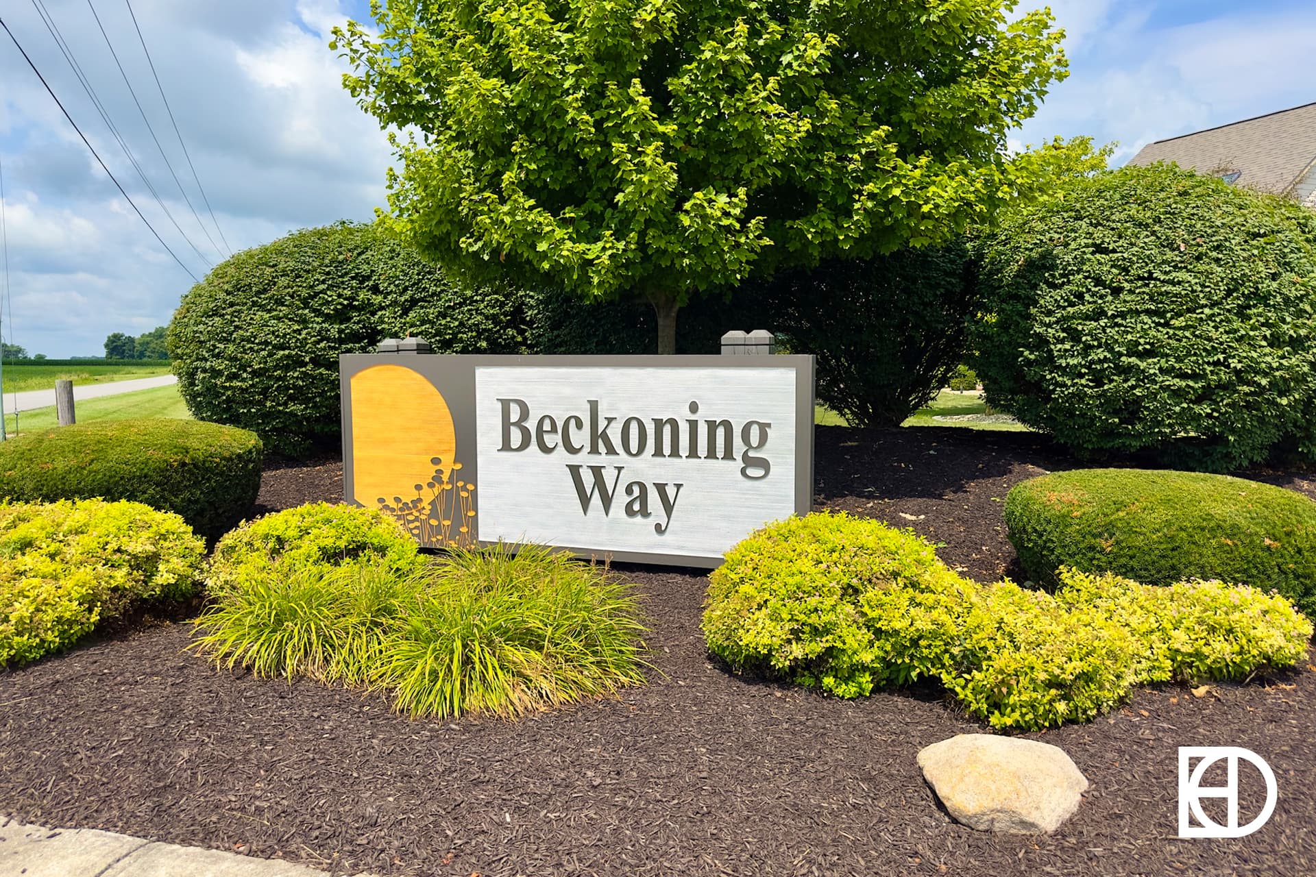 A sign reading “Beckoning Way” stands among neatly trimmed bushes and landscaping mulch, with a tree and a blue sky with clouds in the background.