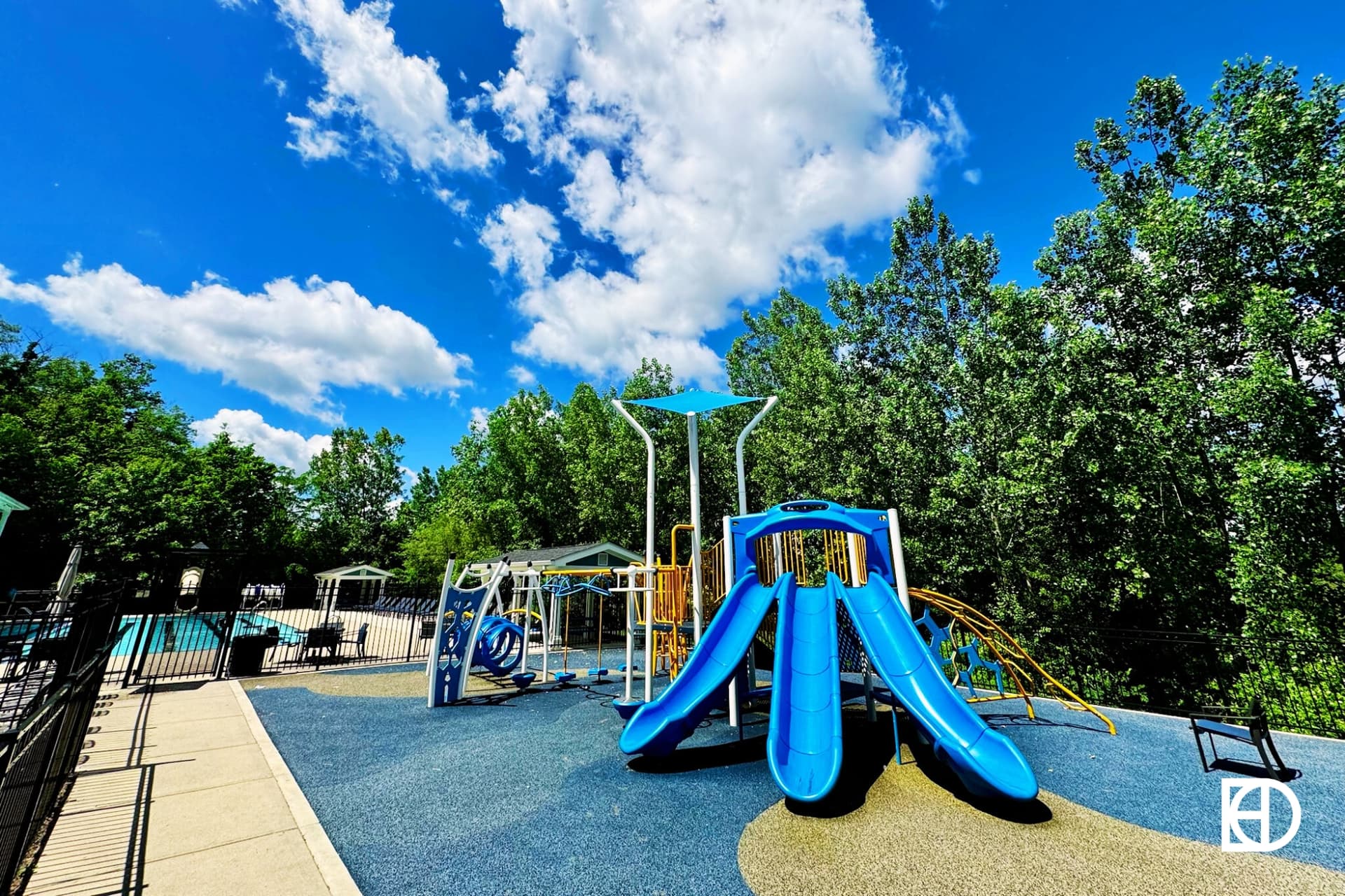 Outdoor playground with blue slides and climbing equipment on a rubber surface, surrounded by trees under a blue sky with scattered clouds.