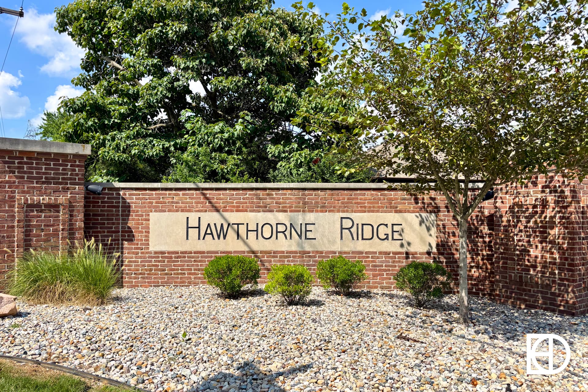A brick entrance sign with Hawthorne Ridge engraved on a stone panel, surrounded by small bushes, rocks, and a tree, against a backdrop of blue sky and greenery.