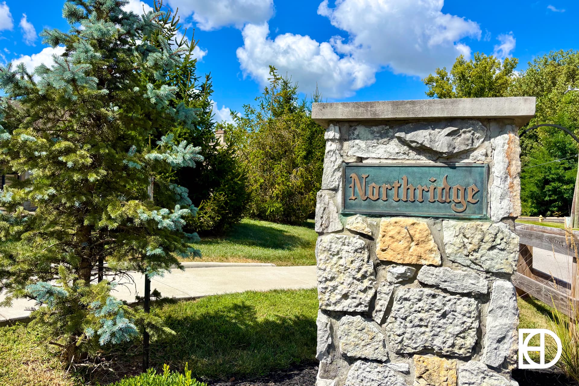 A stone pillar sign reading Northridge stands next to a sidewalk and green trees under a blue sky with scattered white clouds.