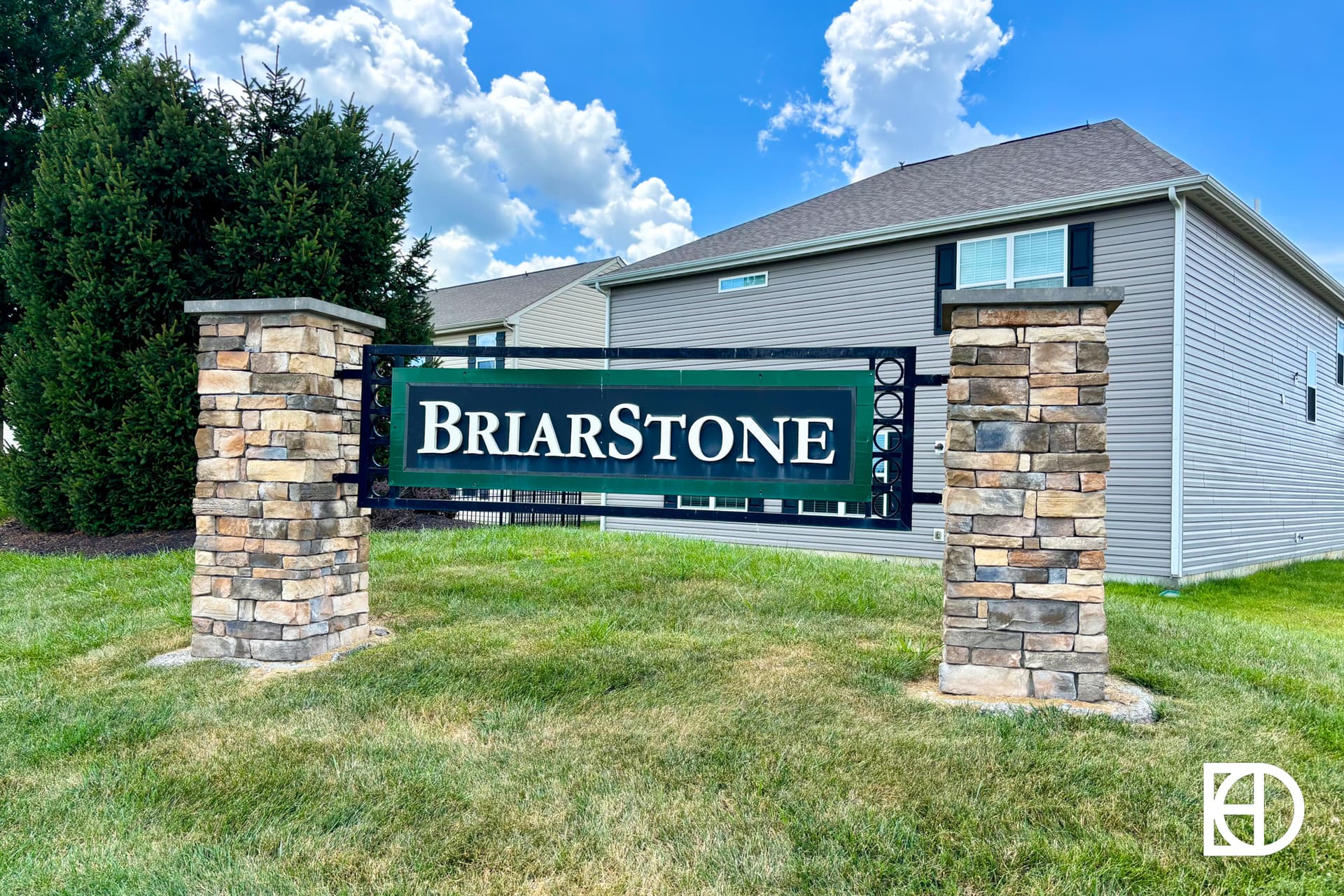 A stone and metal sign reading BriarStone stands on a grassy lawn in front of tan houses, with a blue sky and clouds in the background.