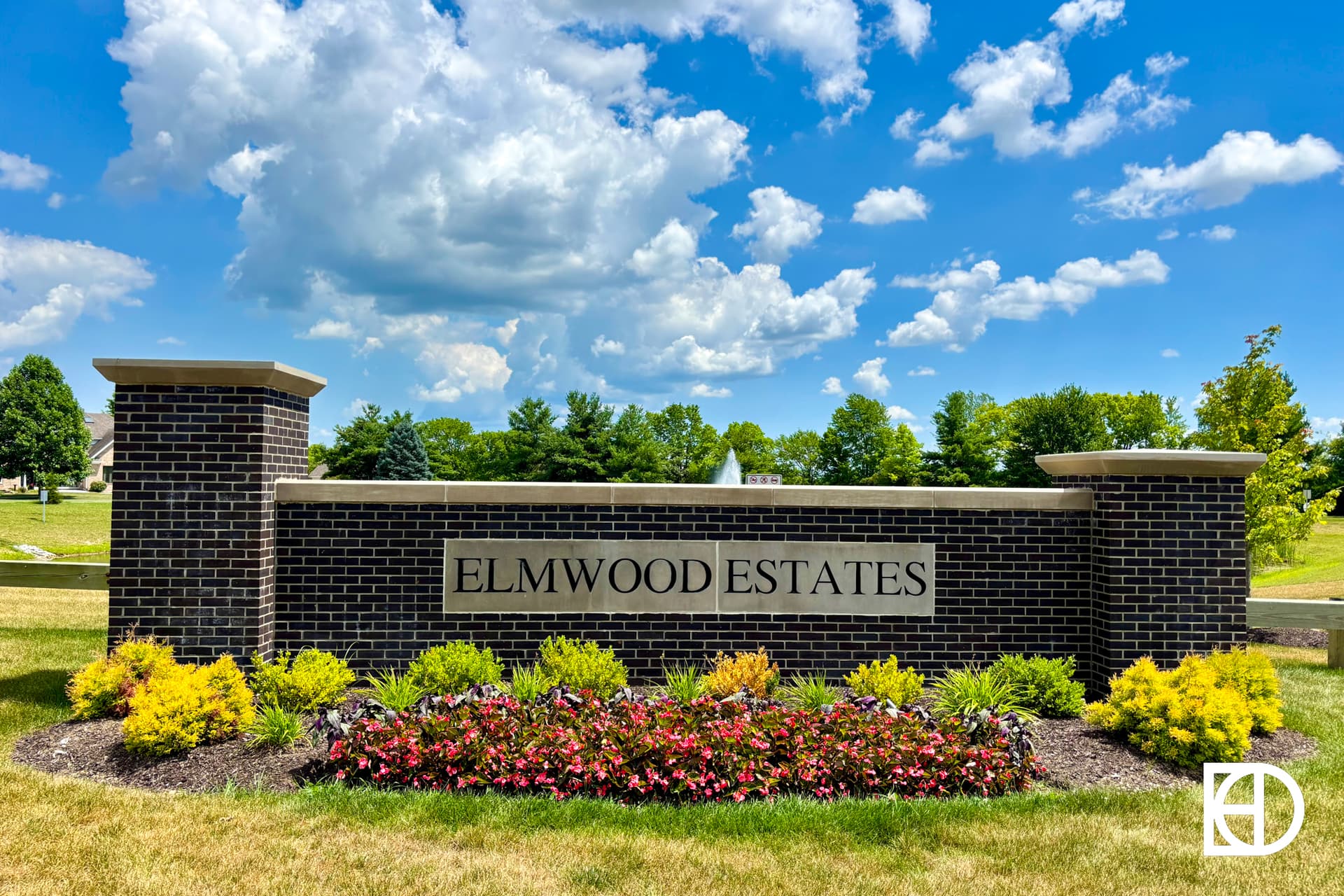 A brick sign reading ELMWOOD ESTATES stands behind colorful flowers and shrubs, with a manicured lawn, trees, and a bright blue sky with scattered clouds in the background.