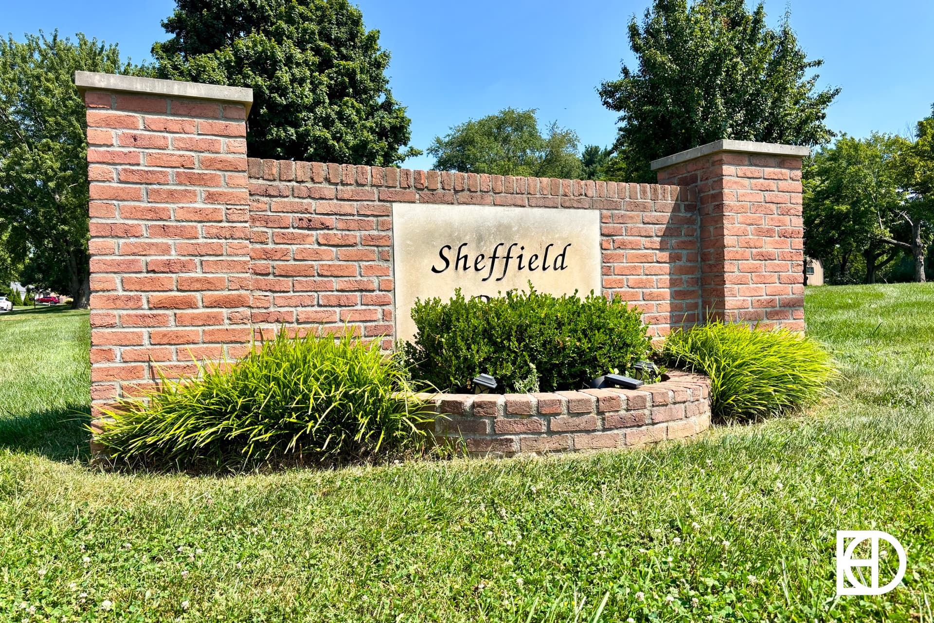 A brick sign with “Sheffield” written on a central stone plaque, surrounded by bushes and green grass, with trees and a clear sky in the background.