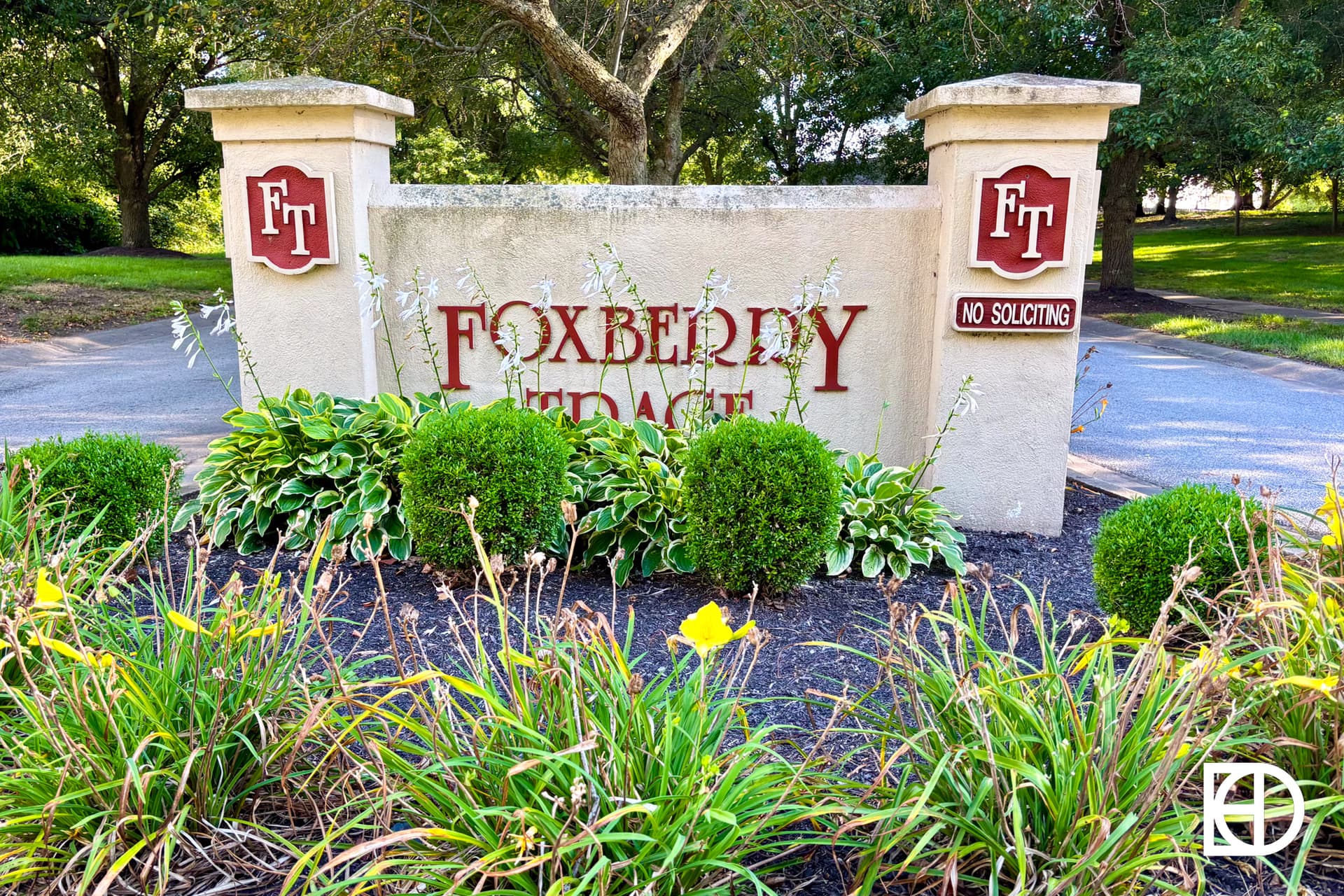A stone entrance sign reads “FOXBERY TERRACE” with the initials “FT” on both sides and a “NO SOLICITING” sign. The sign is surrounded by green shrubs and yellow flowers, with trees and a driveway in the background.