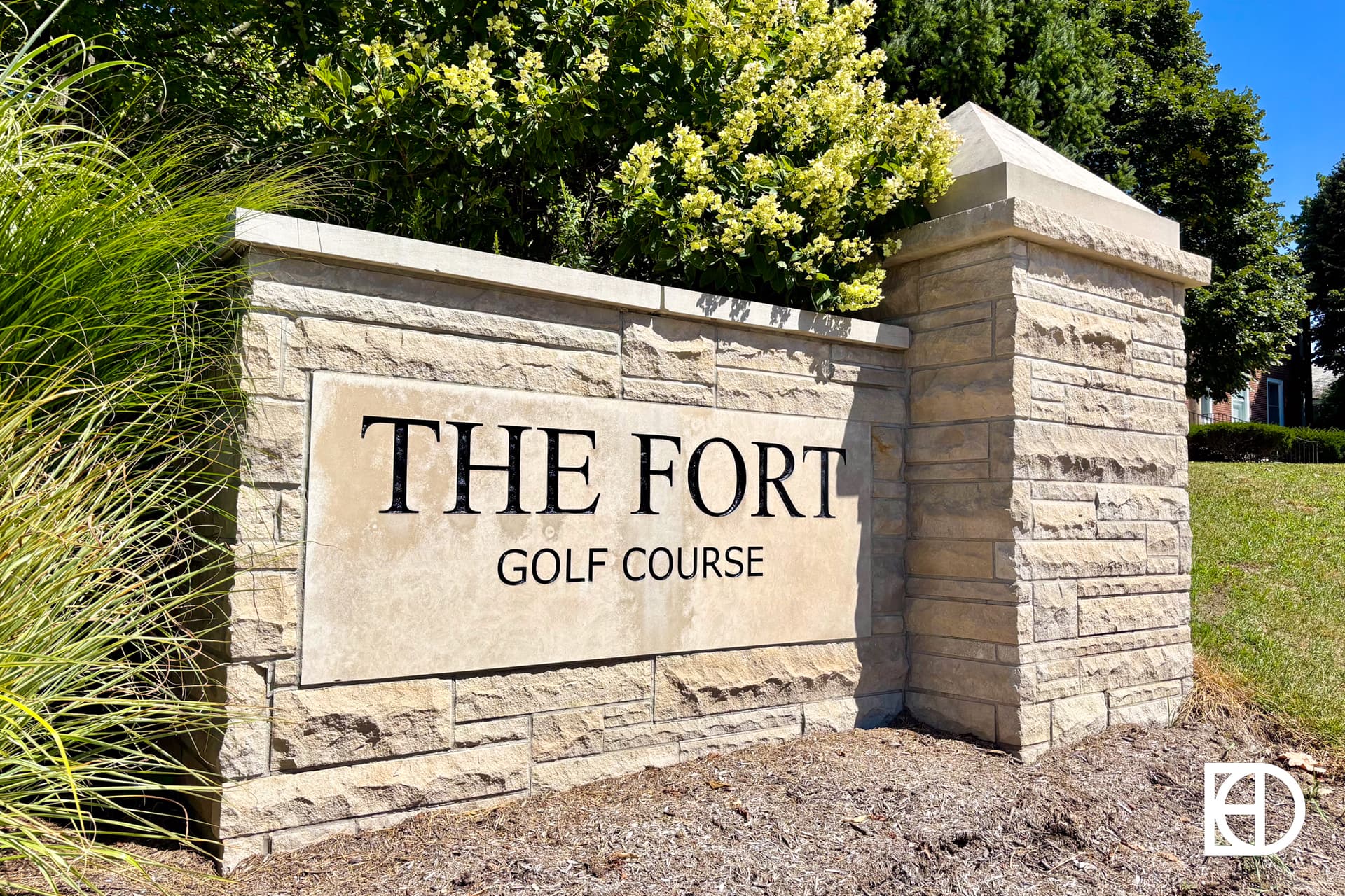 A stone sign with black text reading THE FORT GOLF COURSE is set in a landscaped area with greenery and grass around it. Bright sunlight and blue sky are visible.