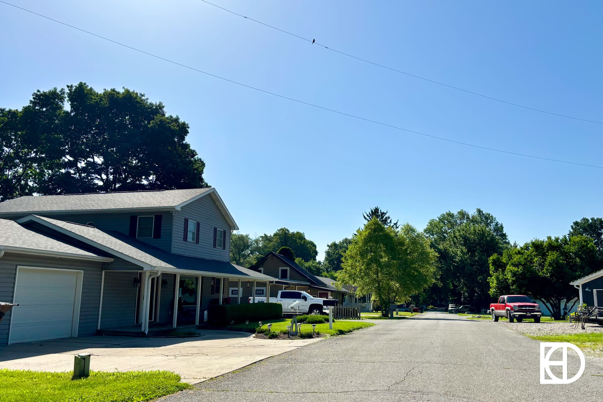 A quiet suburban street with houses, trees, and parked vehicles under a clear blue sky. Sunlight casts shadows, and the neighborhood appears calm and well-kept. The KD logo is in the bottom right corner.