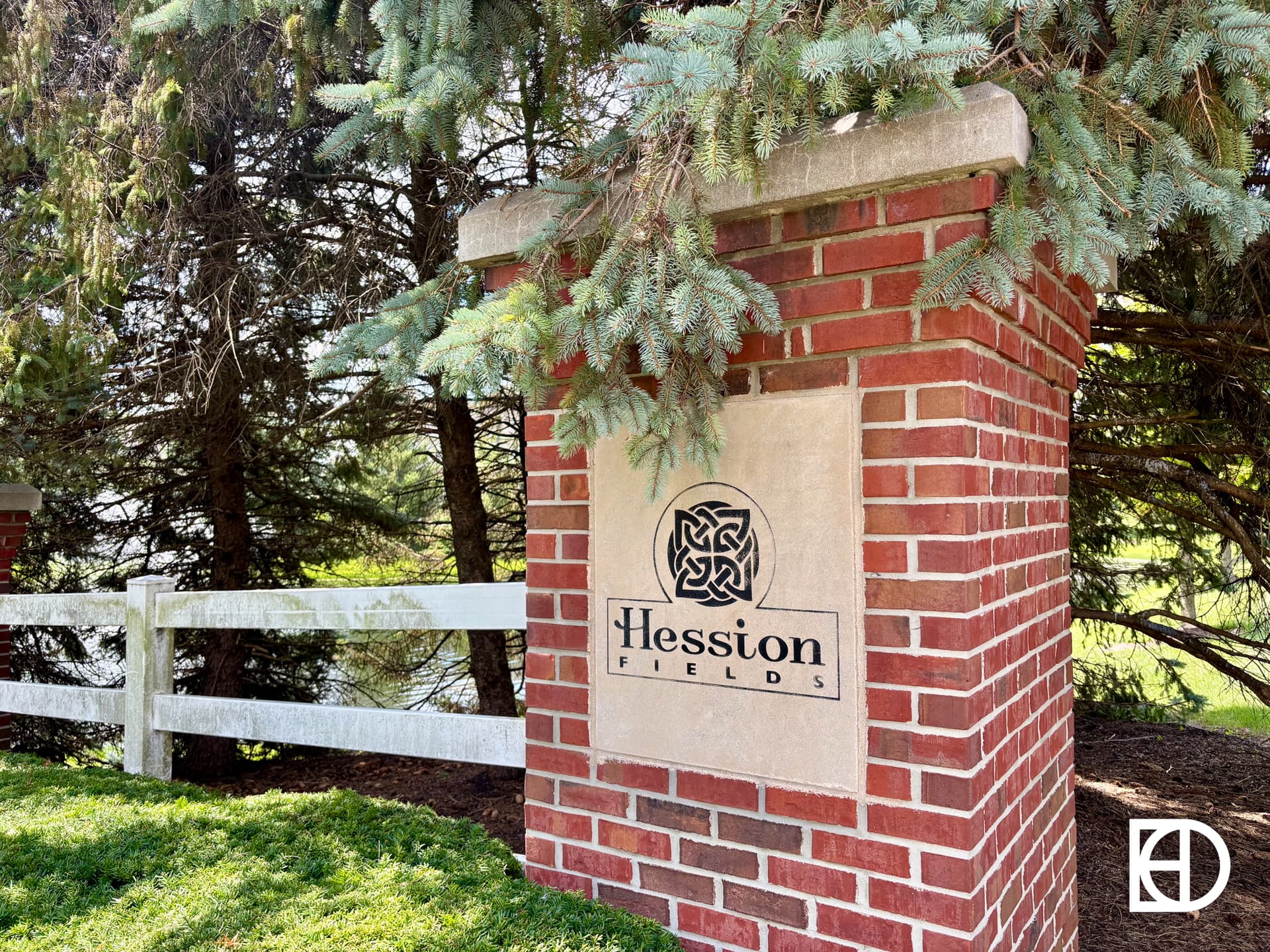 A brick pillar sign reads Hession Fields with a decorative logo, set in front of pine trees and a white wooden fence on a well-maintained lawn.