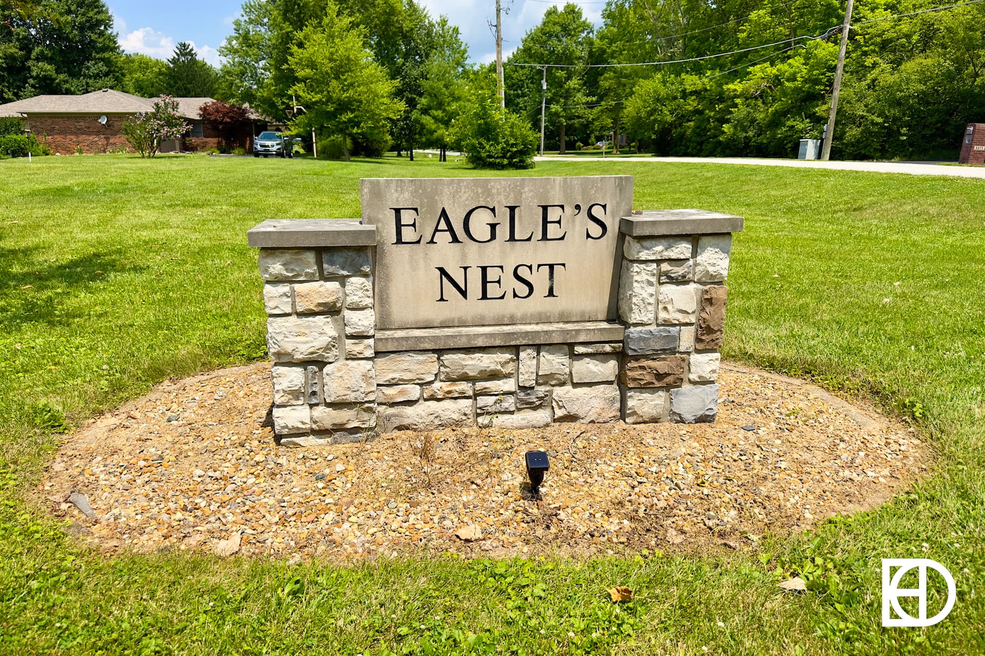 A stone sign reading EAGLES NEST stands on a patch of gravel surrounded by green grass and trees, with a house and street visible in the background.