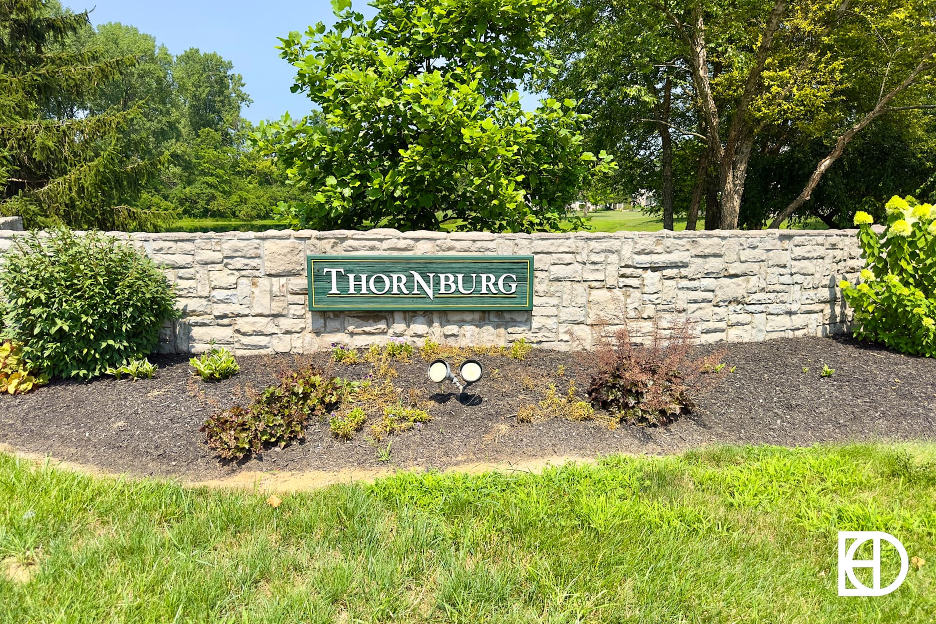 A stone wall with a green sign that reads Thornburg is surrounded by small shrubs and plants on a landscaped mulch bed, with grass and trees in the background.