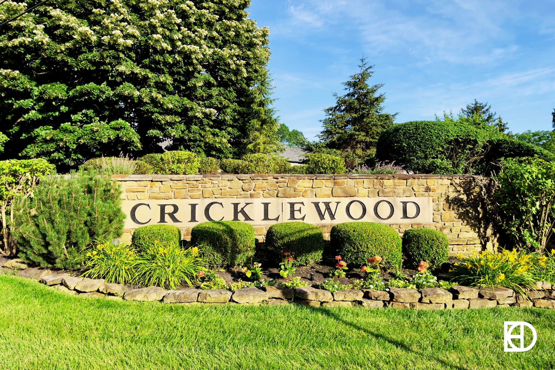 Stone entrance sign to Cricklewood with shrubs and flowers, and trees in the background.