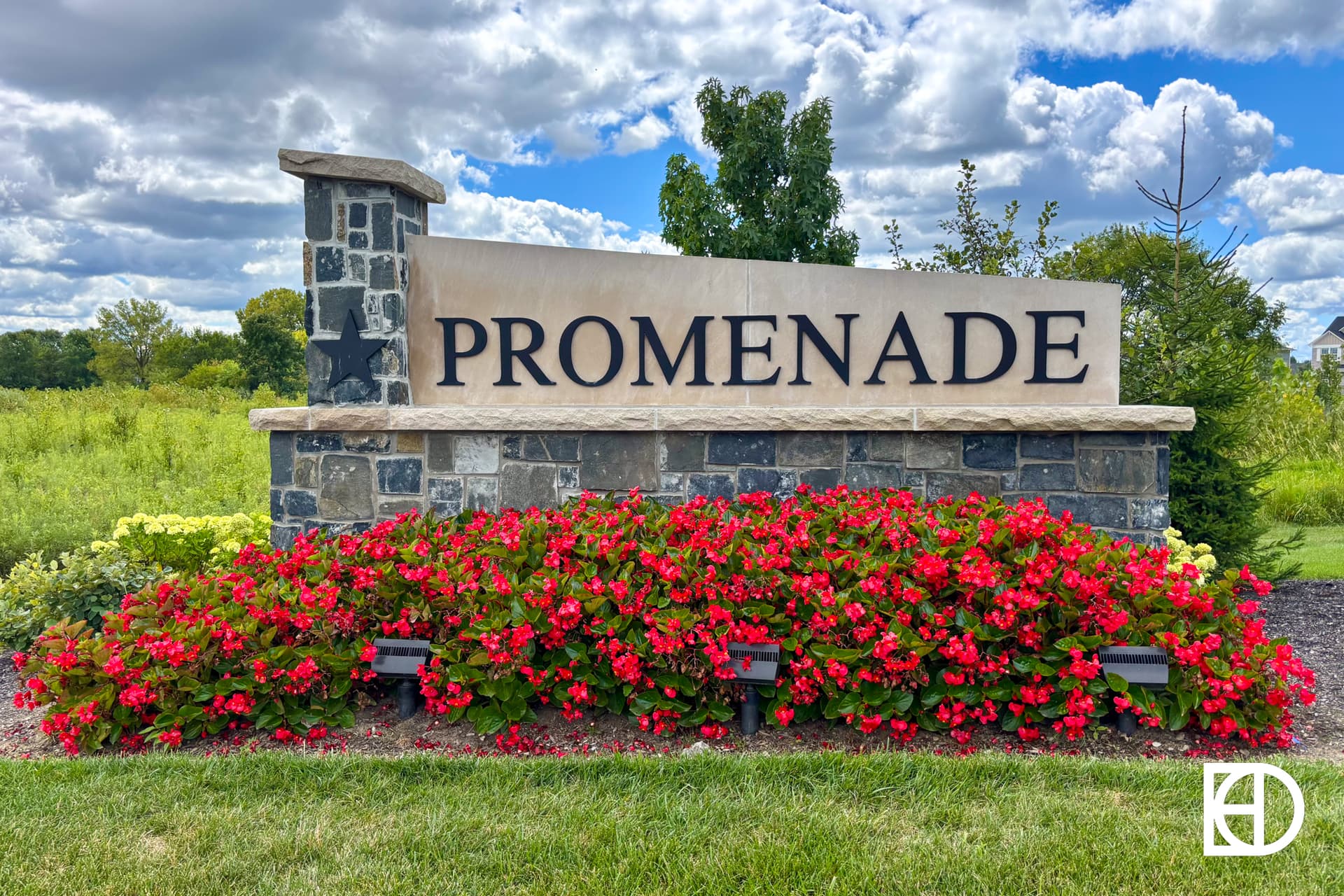 A large stone sign with the word PROMENADE stands behind a bed of vibrant red flowers and greenery, under a partly cloudy blue sky. The KD logo appears in the bottom right corner.